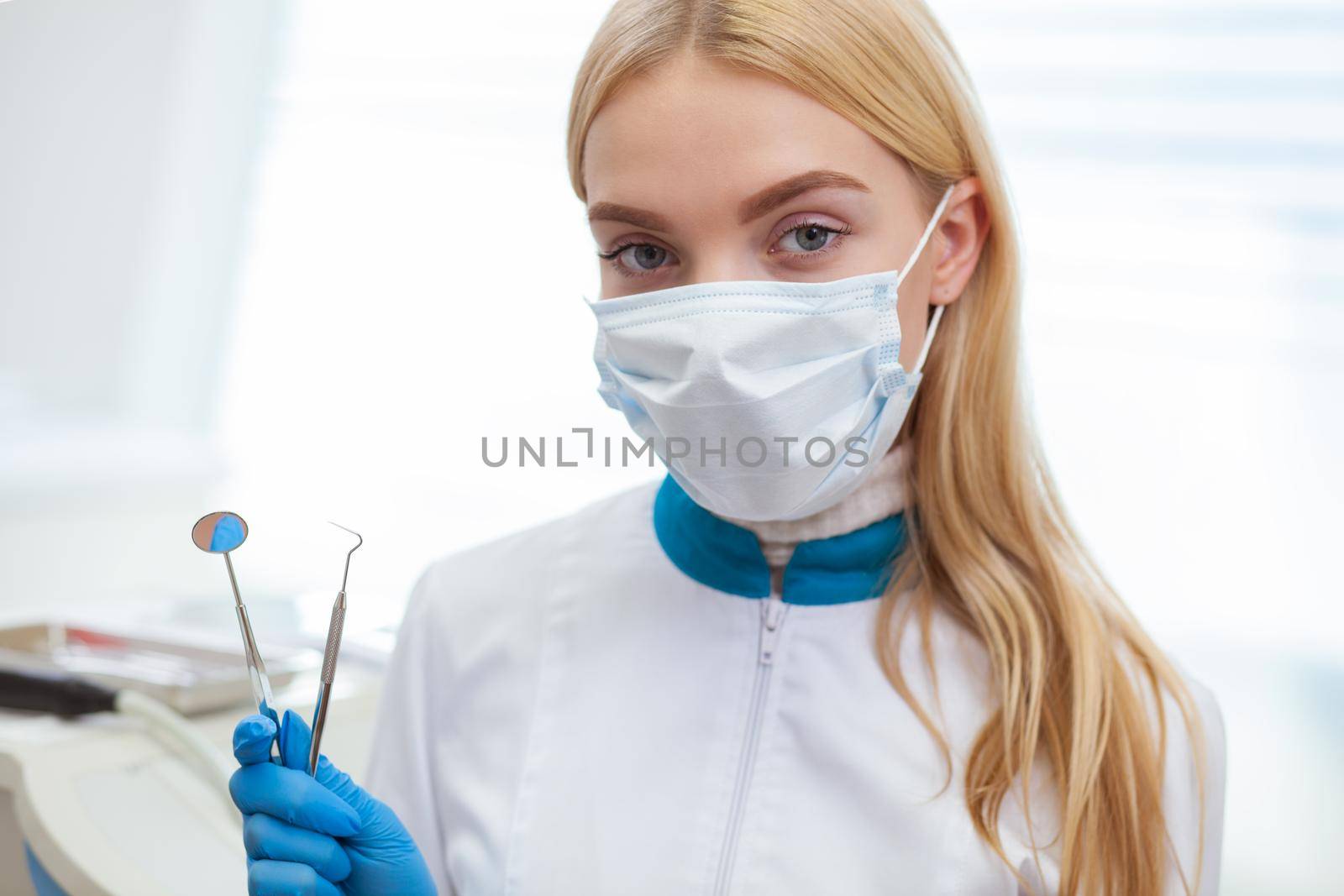 Close up of a young female dentist wearing medical mask holding dental medical tools, looking to the camera. Professional dentist holding oral tools, ready to examine teeth. Smile, teeth care concept