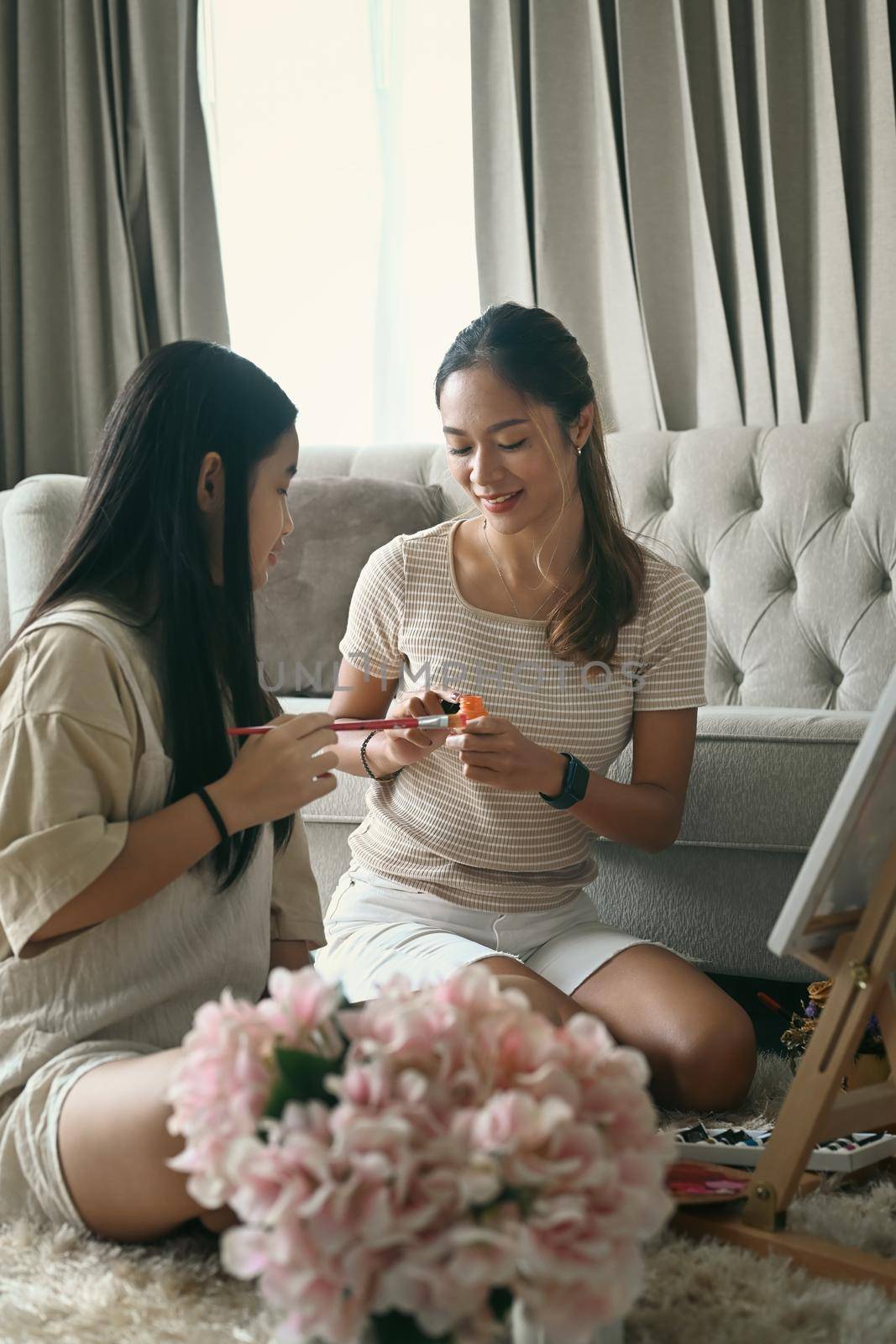 Happy young woman spending leisure time, painting picture with her young sister at home.