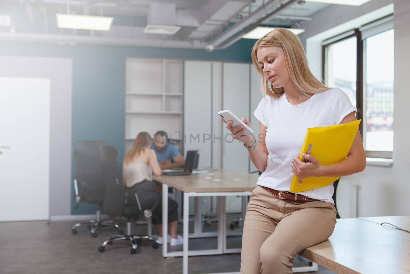 Attractive young businesswoman using smart phone at work, her colleagues on the background