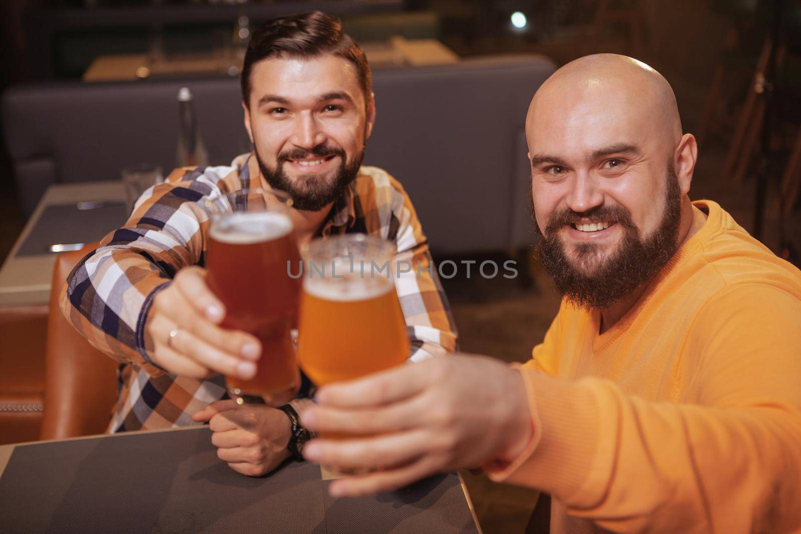 Two cheerful handsome men smiling to the camera, clinking their beer glasses