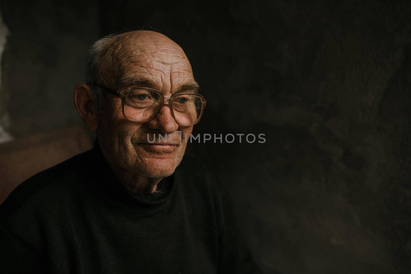 Pensive old man in glasses with gray hair looks away. wrinkles. wisdom. against a dark gray texture wall. bald head. in a knitted sweater. Portrait.