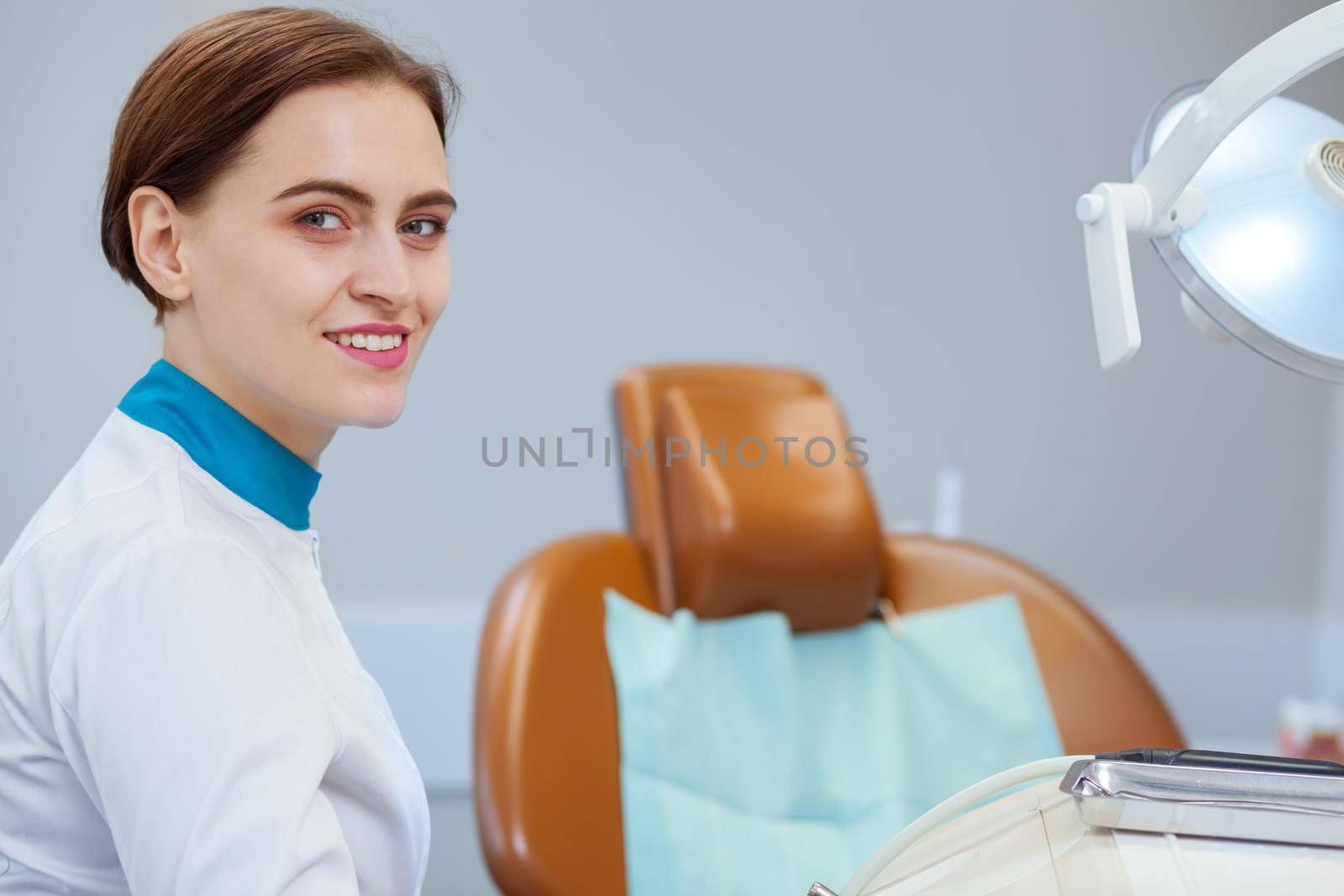 Attractive female doctor smiling to the camera over her shoulder. Professional dentist waiting for her patient at the dental clinic, smiling joyfully, copy space. Feminism, success, women concept