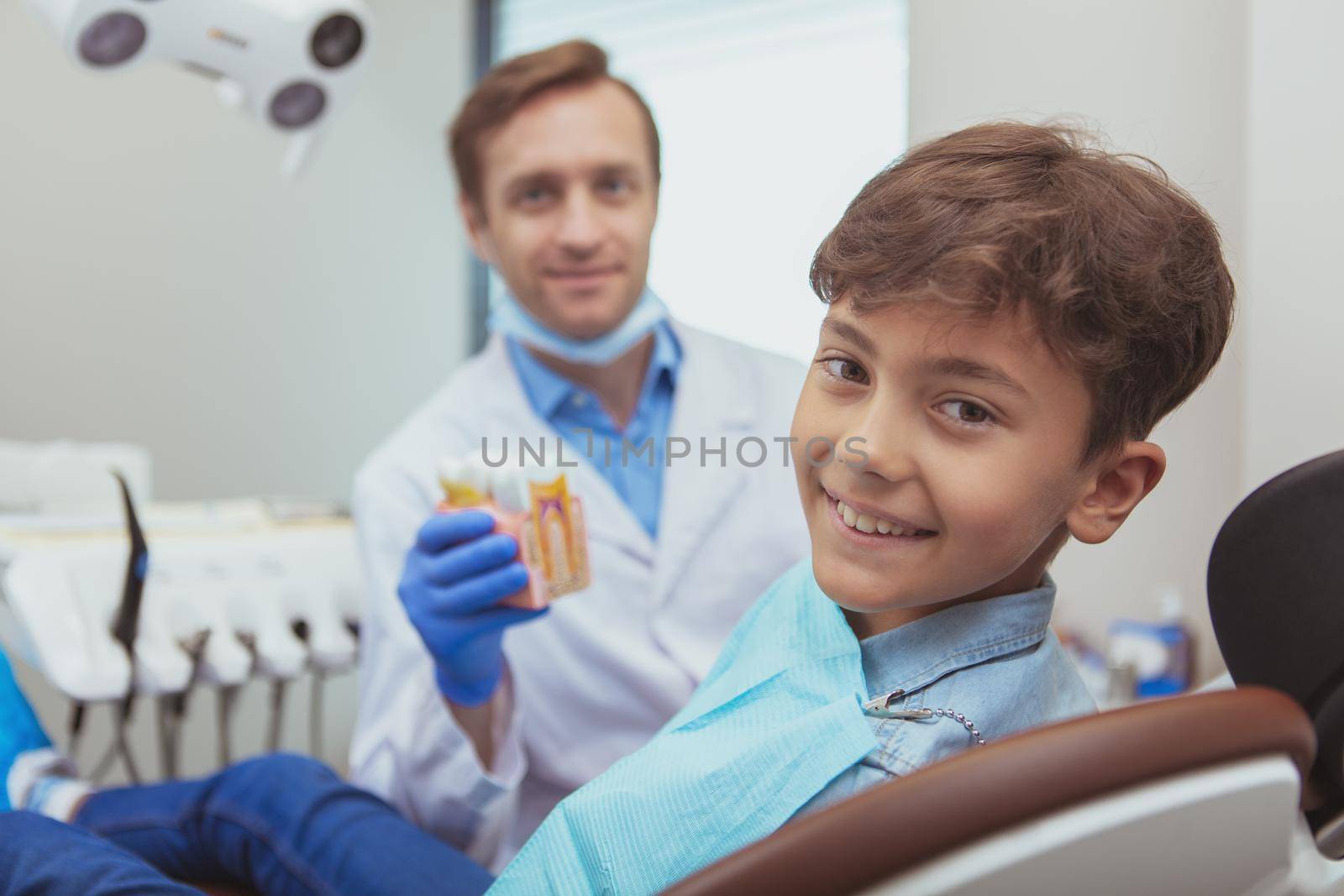 Healthy teeth, children concept. Adorable happy boy smiling to the camera while sitting in a dentist chair, dentist smiling on the background