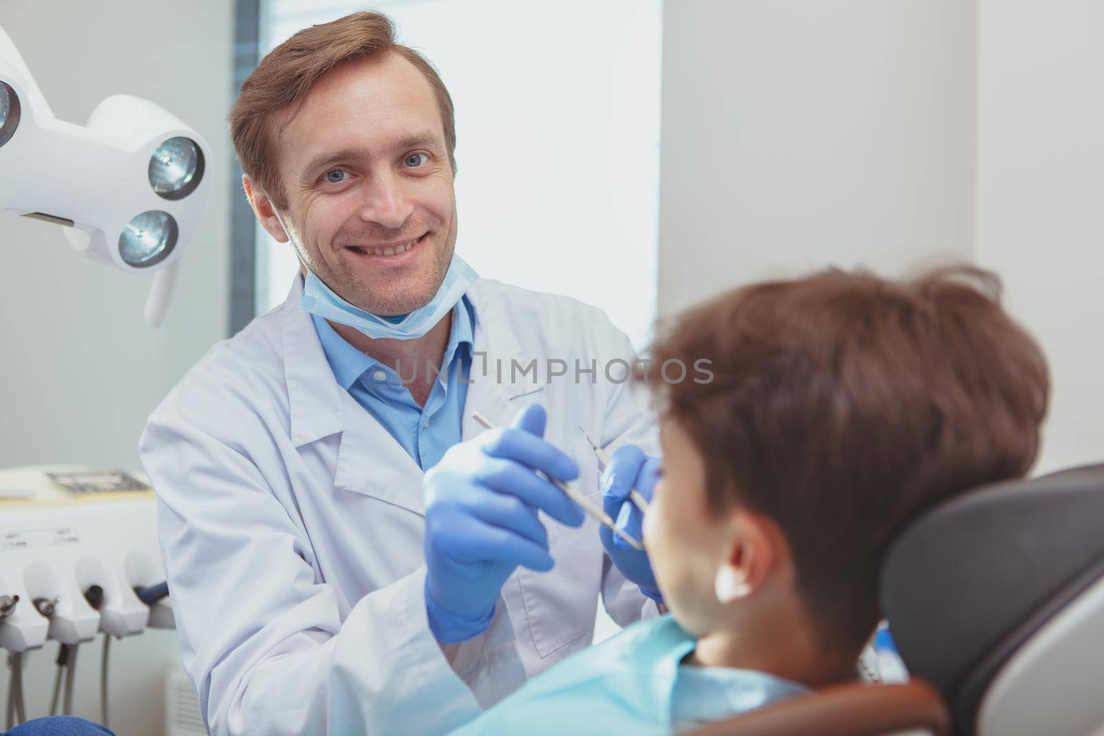 Experienced dentist concept. Handsome mature male dentist smiling joyfully to the camera, working at his clinic, examining teeth of his little patient