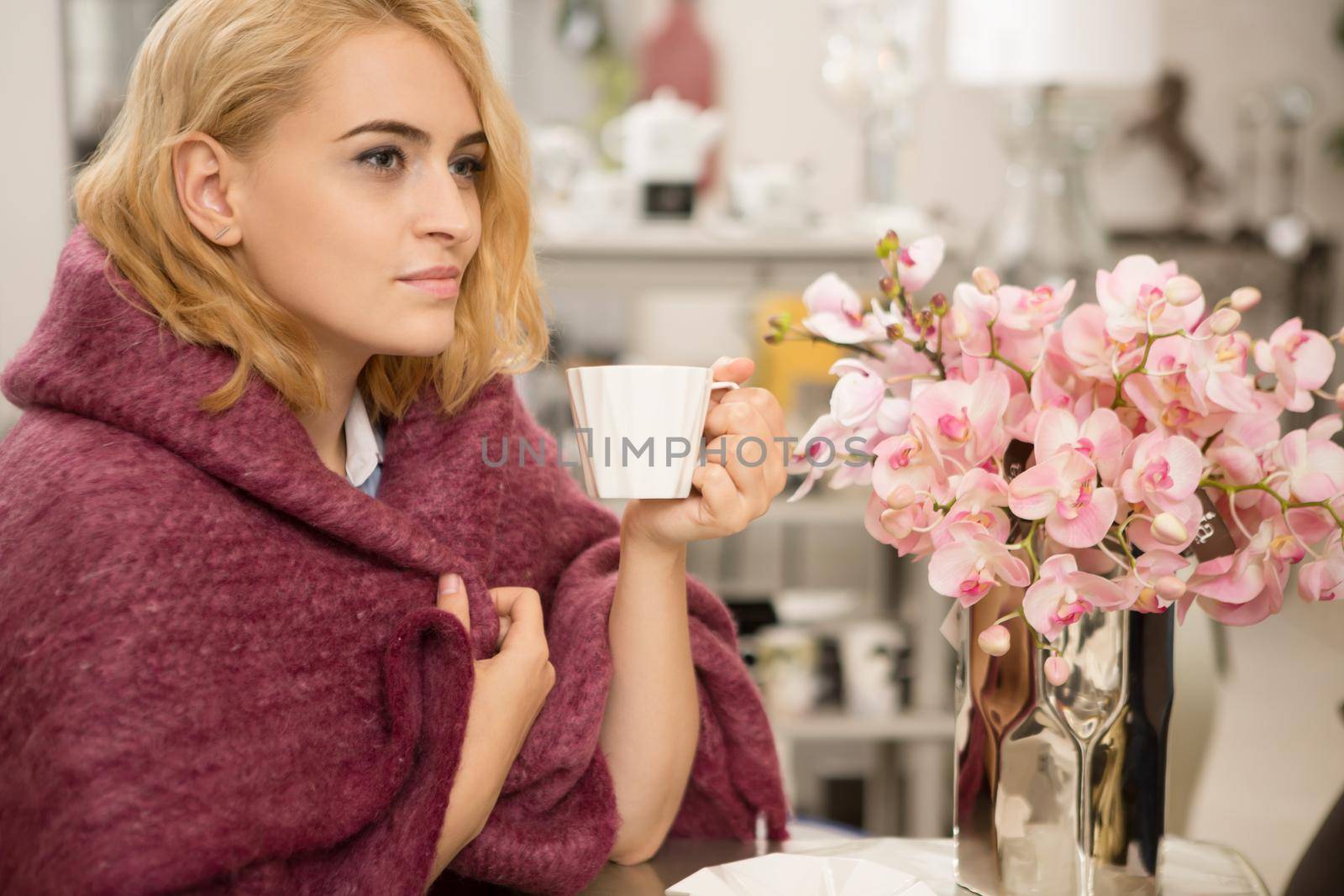 Attractive blonde young woman enjoying cup of tea or coffee sitting wrapped in a woolen warm blanket relaxing at home on a weekend copyspace lifestyle relaxation harmony femininity beauty leisure