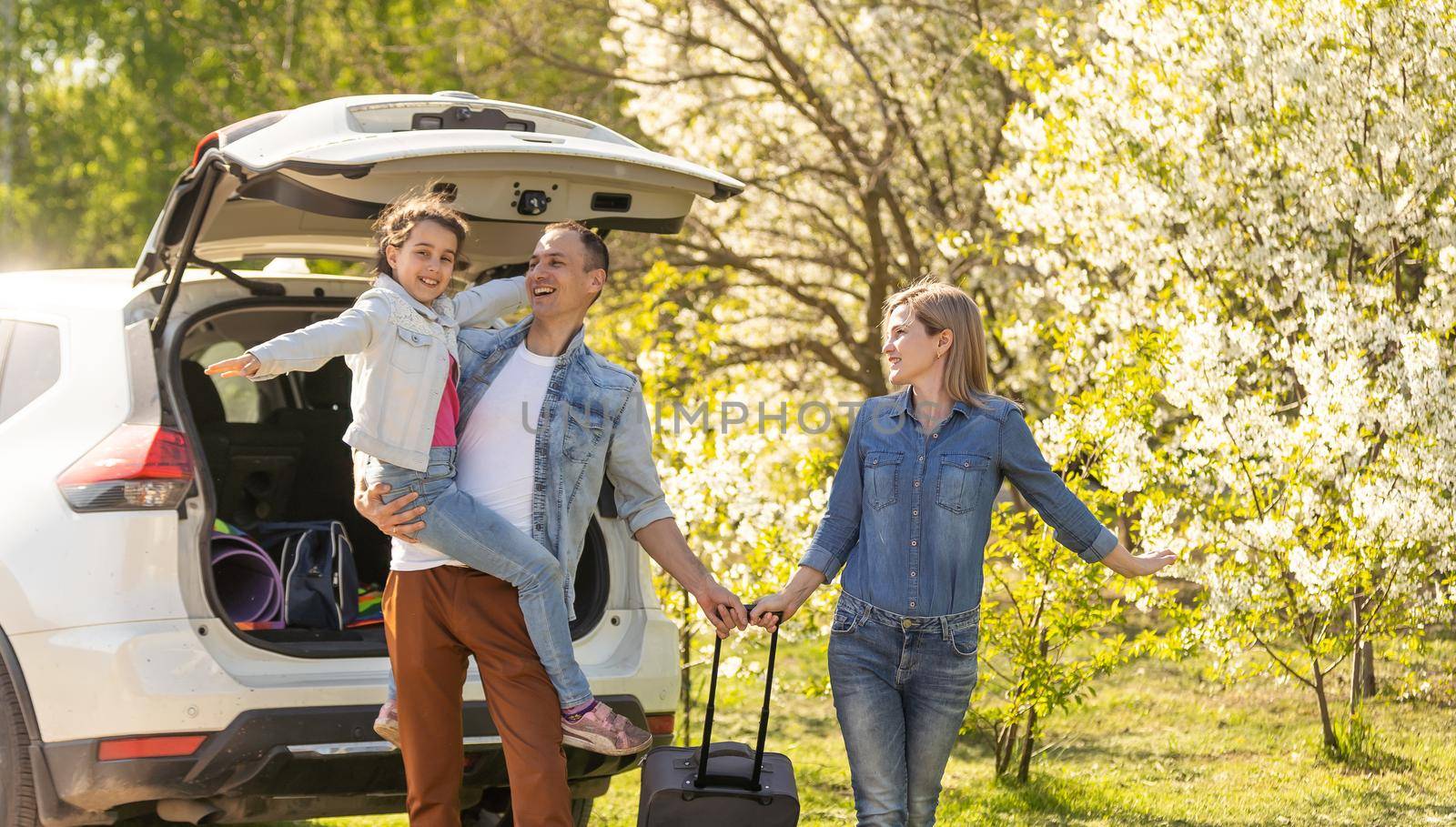 Happy family standing together near a car with open trunk enjoying view of rural landscape nature. Parents and their kid leaning on vehicle luggage compartment. Weekend travel and holidays concept. by Andelov13