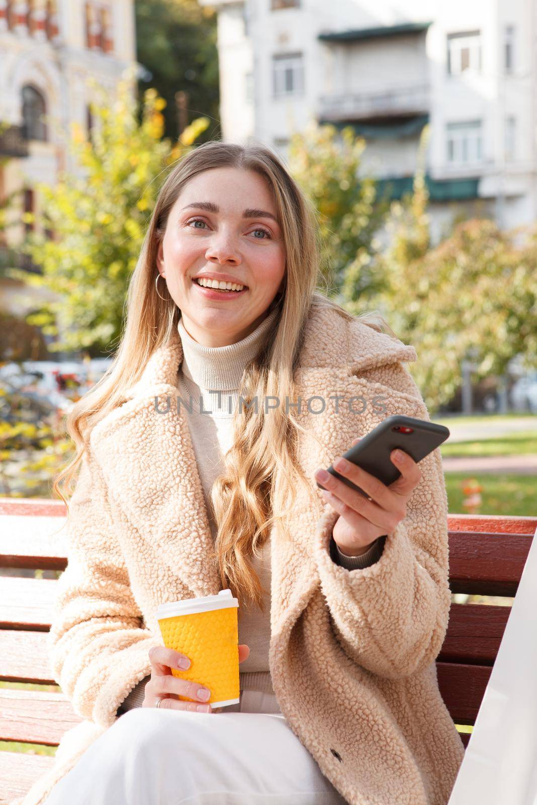 Vertical shot of a beautiful happy woman having coffee in the park in autumn, using her smart phone