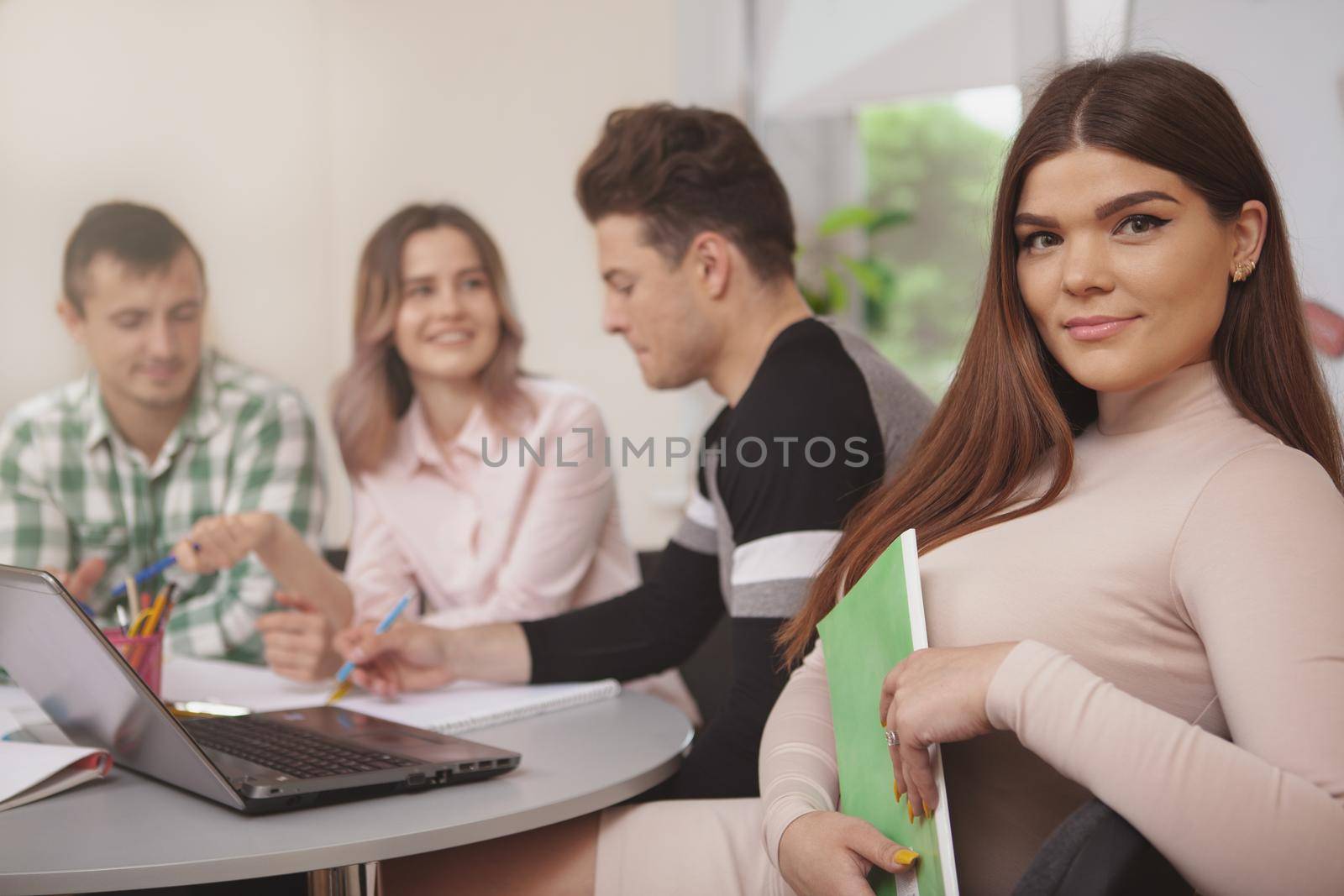 Beautiful young female student smiling to the camera, her classmates working on the background, copy space. Cheerful lovely woman enjoying working on a project with her college friends