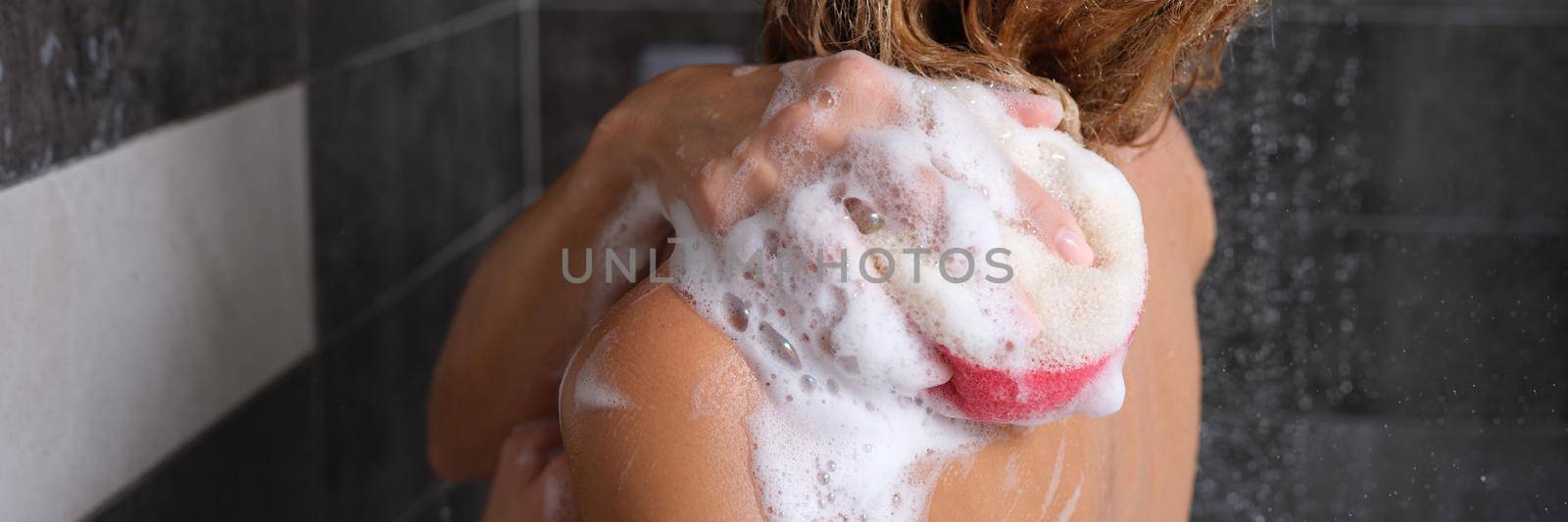 A woman in the shower with a soapy sponge washes her body, view from the back. Shower gel foam on a washcloth, personal hygiene