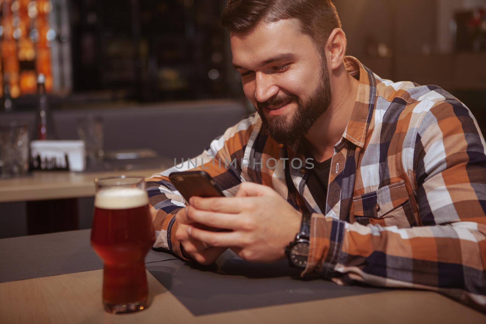 Happy handsome man texting on his smart phone at beer pub. Attractive cheerful man using his phone while drinking beer at the bar