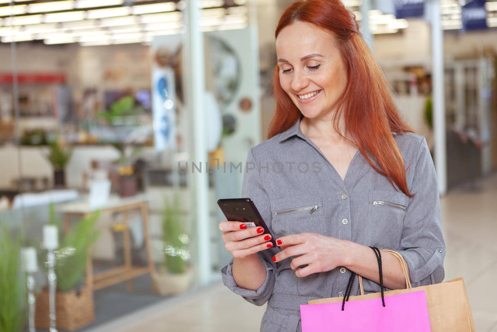 Cheerful woman using her smart phone at the shopping mall, copy space. Attractive female customer walking with shopping bags at the mall, browsing online on her phone