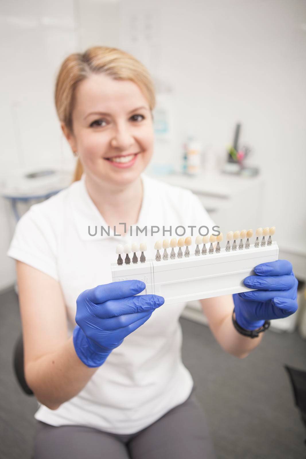 Vertical shot of a cheerful female dentist holding out teeth whitening shade guide tot he camera