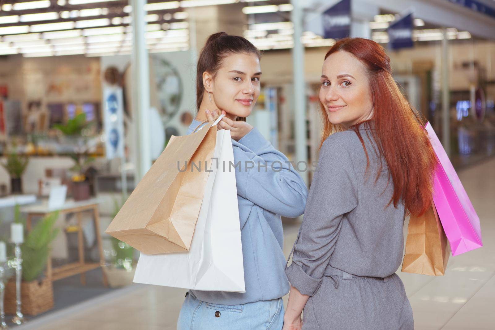 Two charming female friends smiling to the camera over their shoulders while walking at the mall with shopping bags