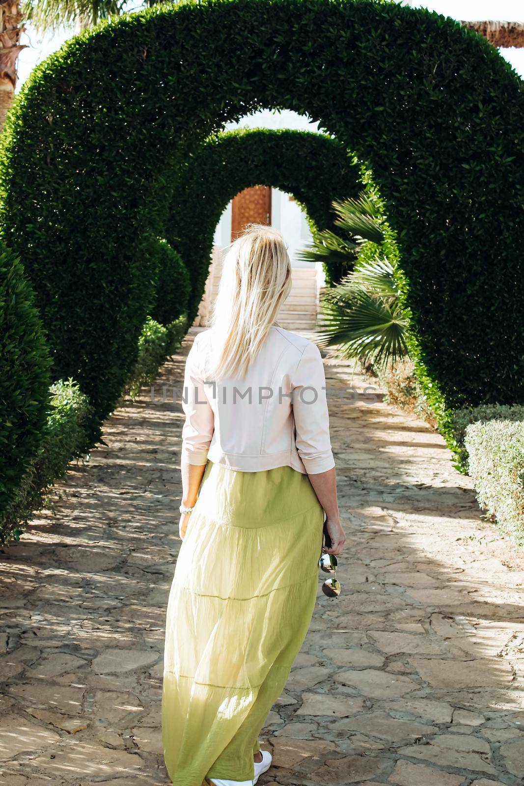 A woman goes forward. View from the back of a walking woman along the alley of arches from green plants