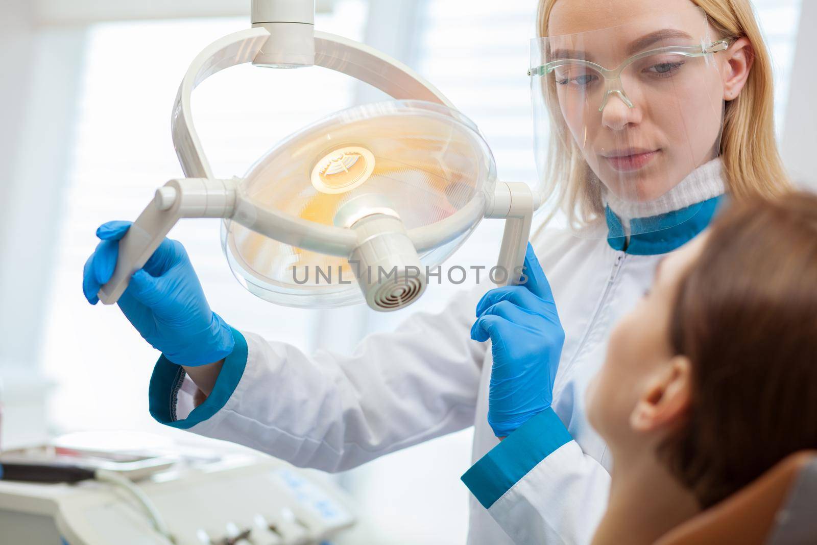 Attractive female dentist adjusting dental lamp, preparing to examine teeth of her patient. Professional dentist working at her clinic. Woman visiting dentist for teeth examination. Profession concept