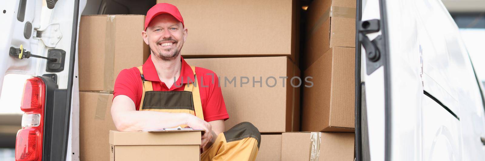 Joyful male courier is holding a parcel while sitting in the truck. Address express delivery of goods during lockdown