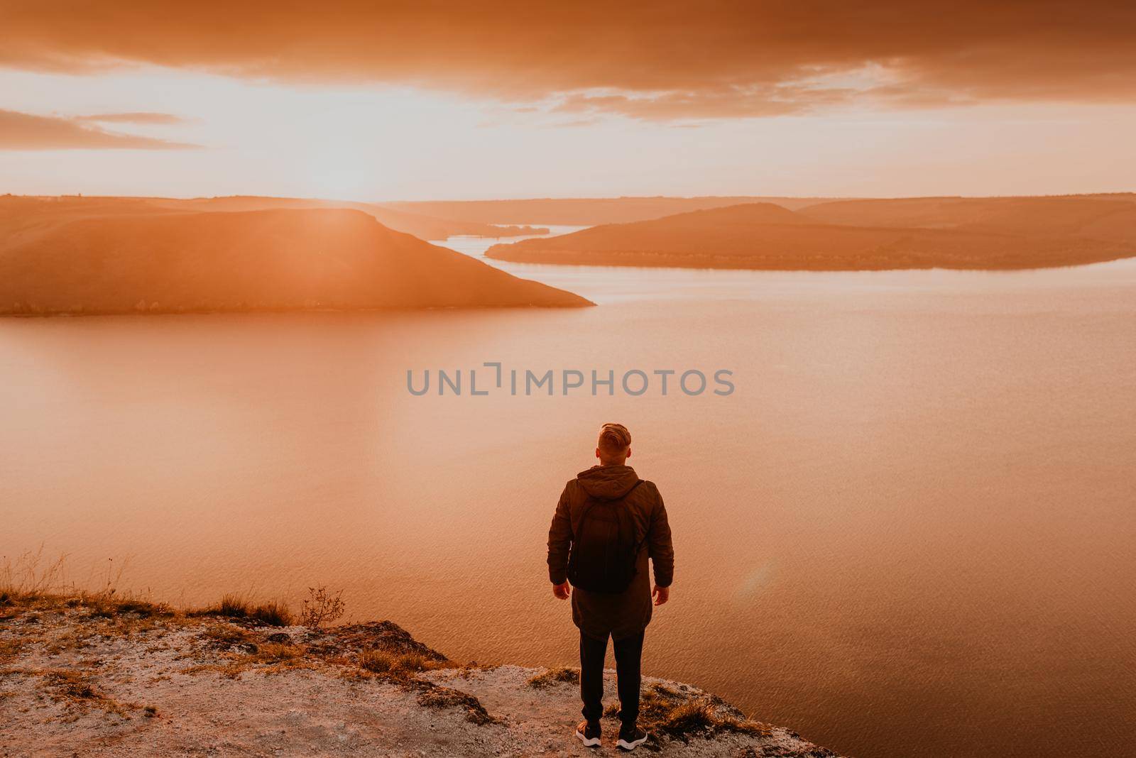 man in casual clothes stands on mountain. fiery orange sunset sunset rays through clouds. panoramic view from cliff to river lake sea in distance silhouettes islands