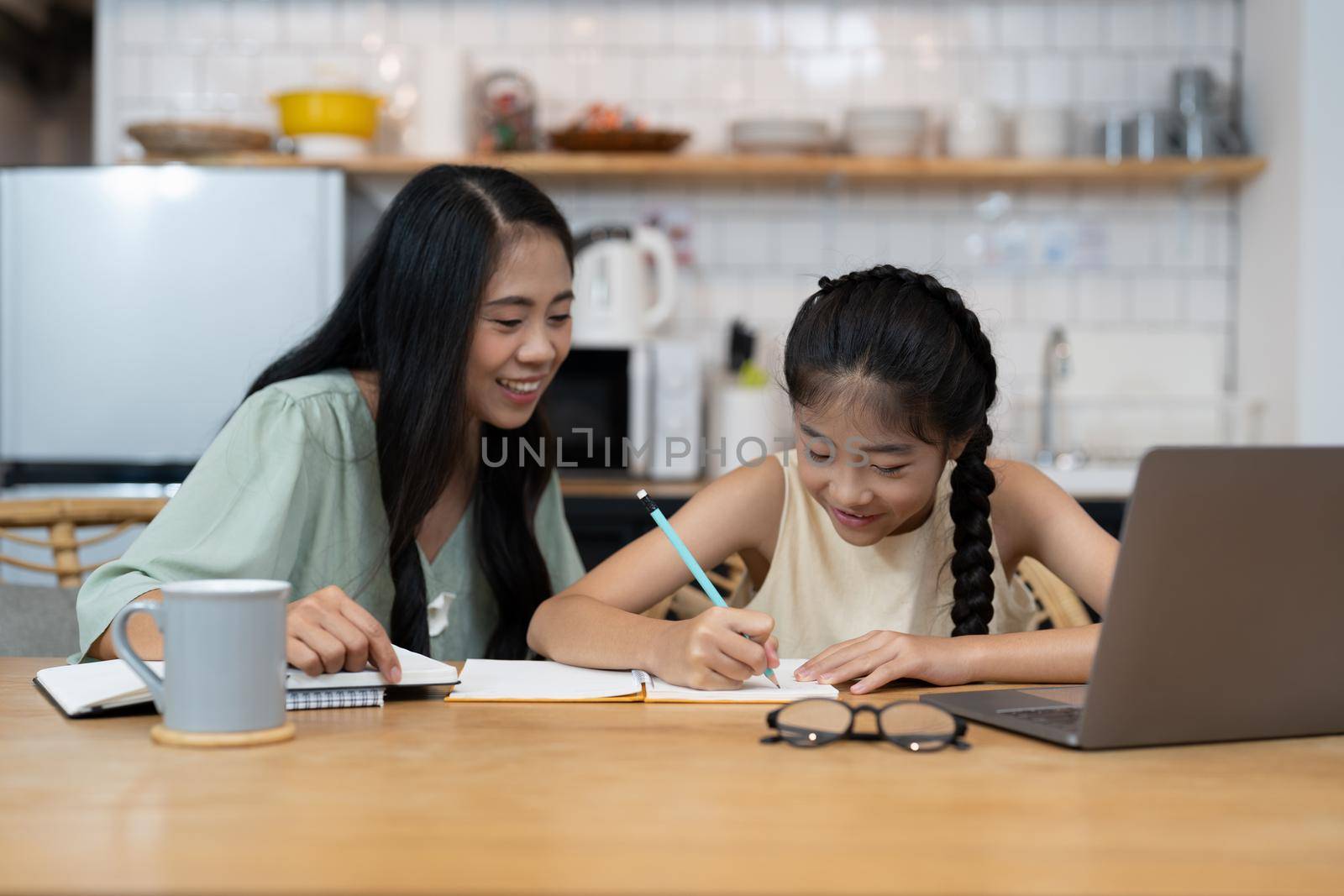 Mother and asian kid little girl learning and looking at laptop computer making homework studying knowledge with online education e-learning system. children video conference with teacher tutor at home.