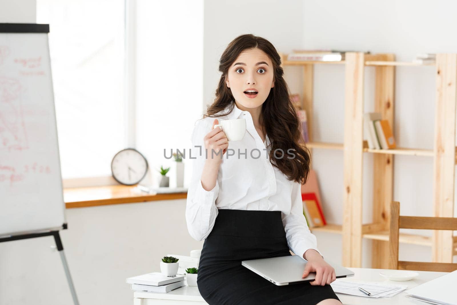 Attractive smiling business woman sitting at office desk, holding a cup of coffee, she is relaxing and happy.