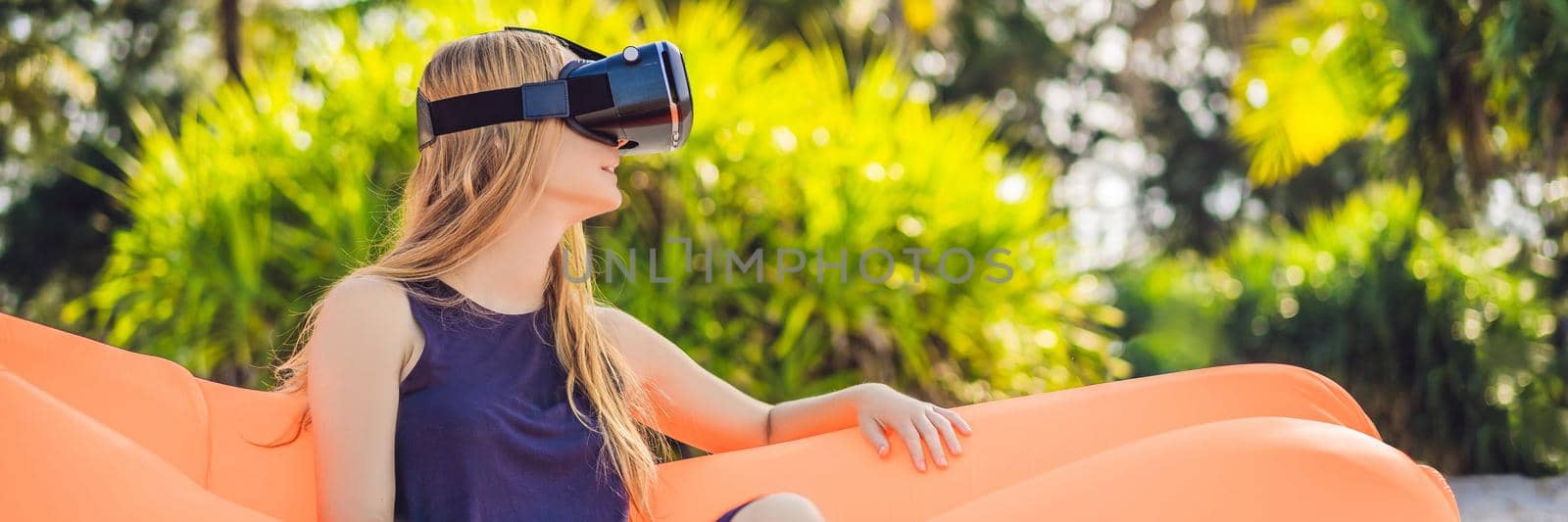 BANNER, LONG FORMAT Summer lifestyle portrait of pretty girl sitting on the orange inflatable sofa and uses virtual reality headset on the beach of tropical island. Relaxing and enjoying life on air bed by galitskaya