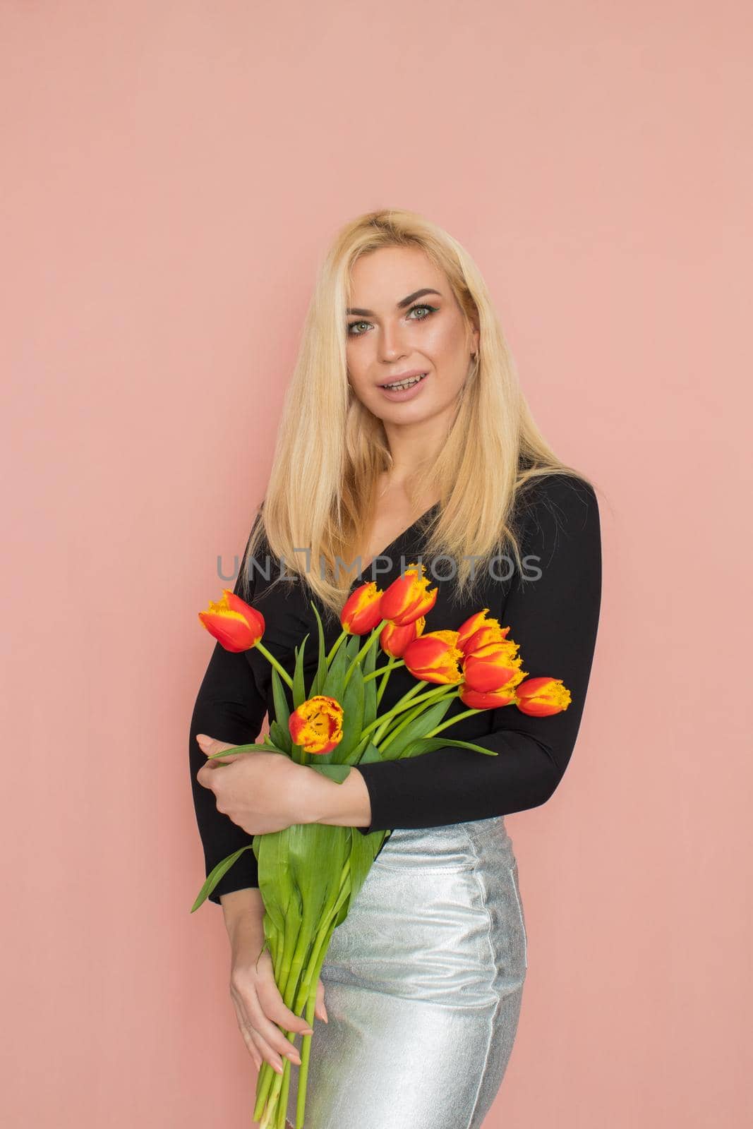 Fashion model woman in fashionable clothes on pink background. Wearing stylish clothing, black blouse, silver skirt. Posing in studio. Holding red tulips in her hands