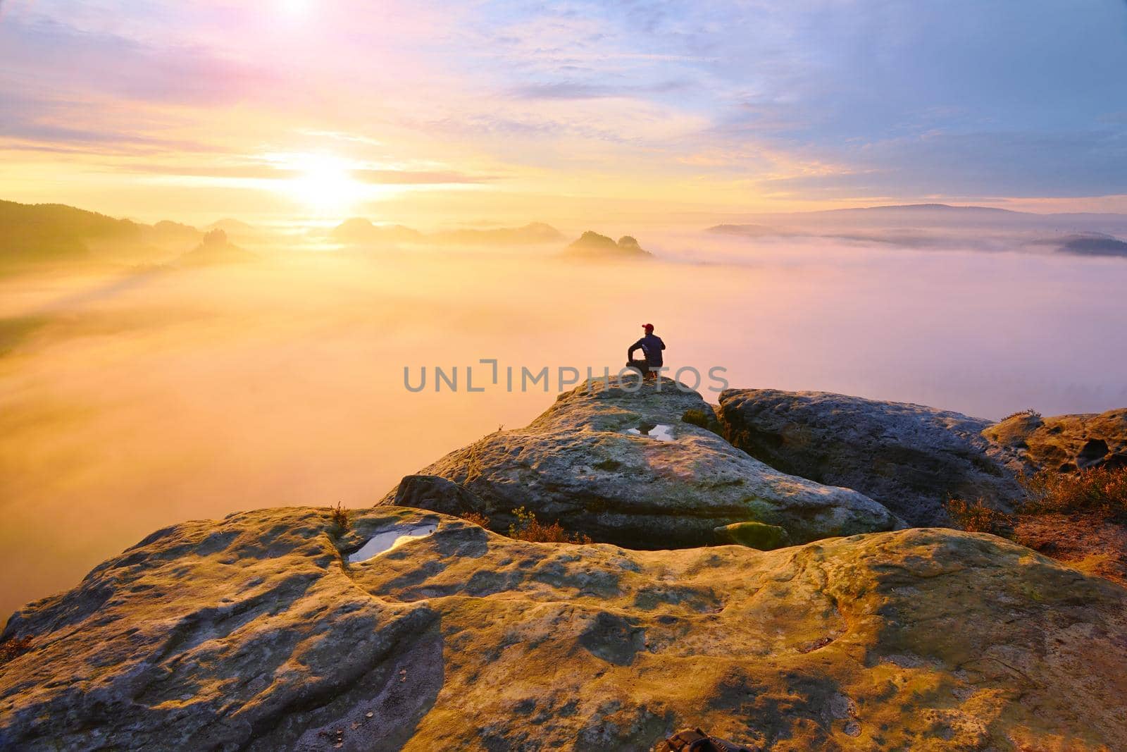Hiker in squatting position on peak of rock and watching into colorful mist and fog in morning valley. by rdonar2