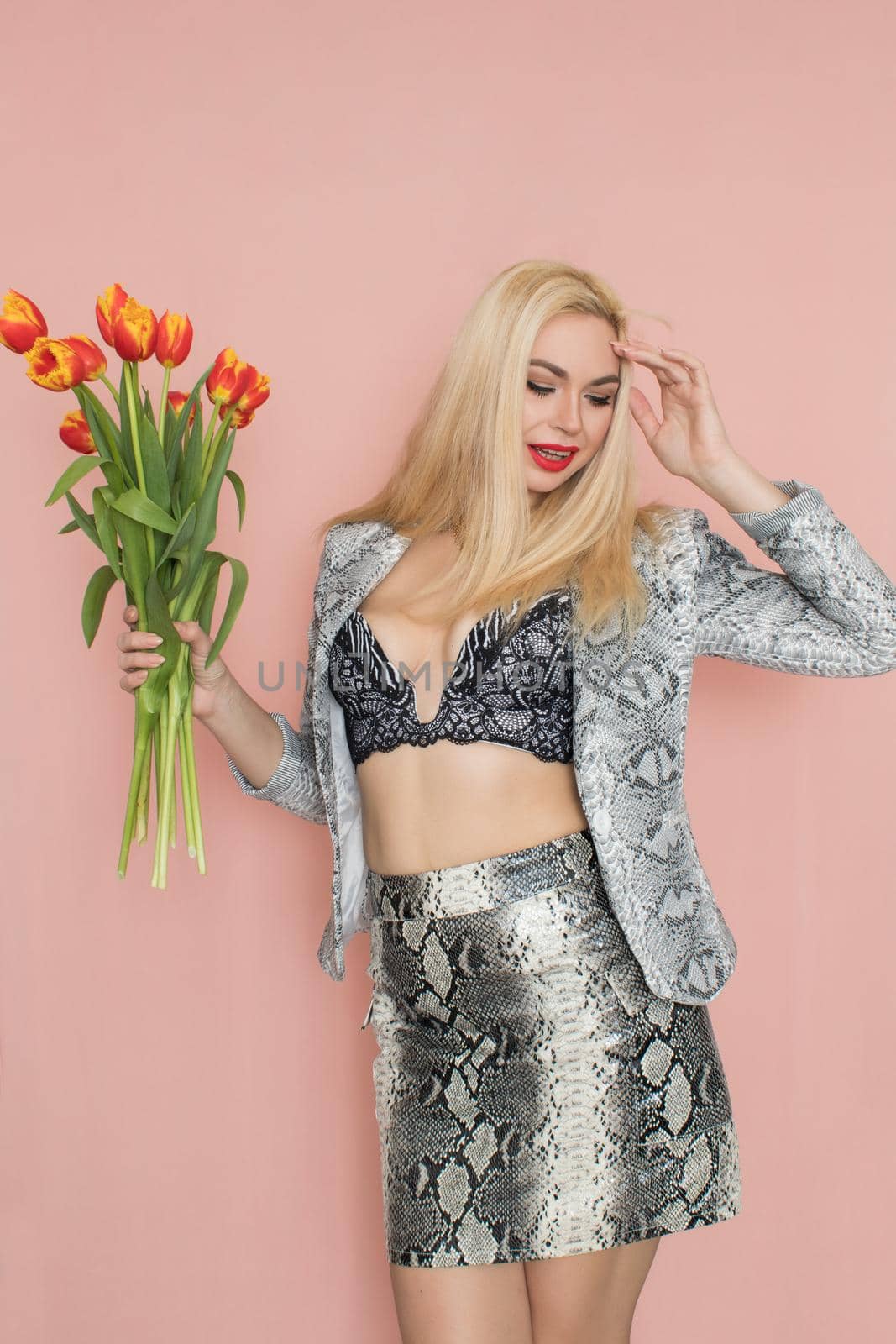 Fashion photo of a beautiful elegant young woman in pretty snake suit, jacket blazer, top, skirt, massive chain around the neck posing on pink background. Studio shot. Bouquet orange tulips holding in hands.