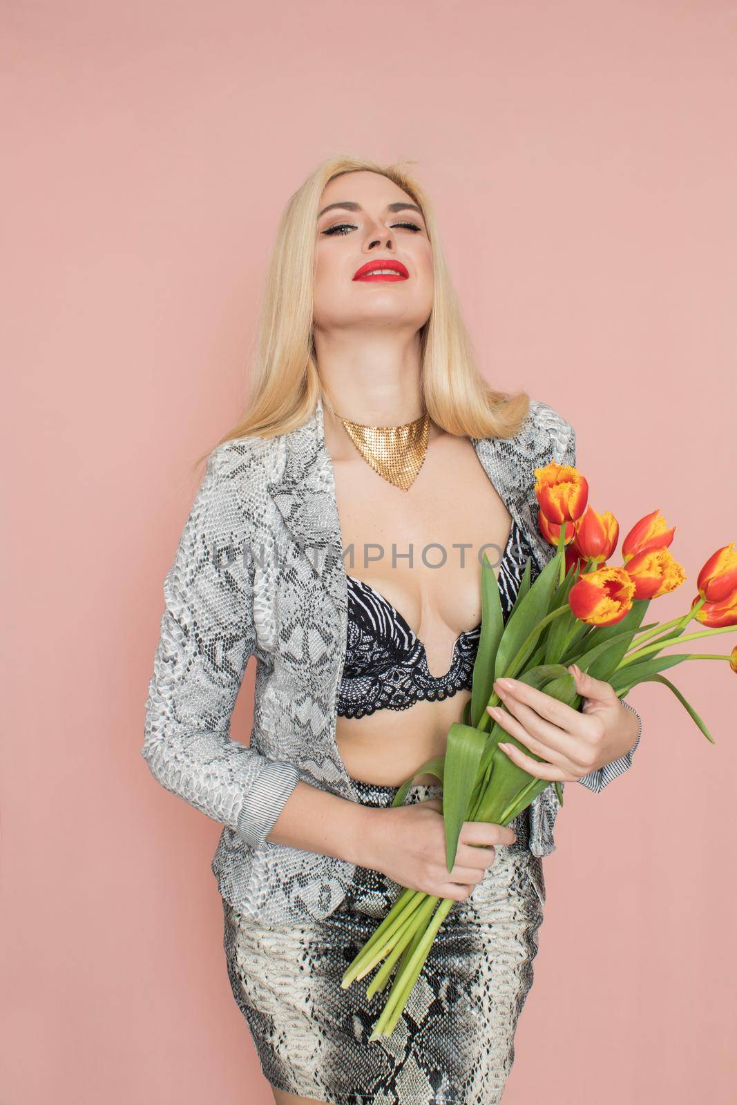 Fashion photo of a beautiful elegant young woman in pretty snake suit, jacket blazer, top, skirt, massive chain around the neck posing on pink background. Studio shot. Bouquet orange tulips holding in hands.