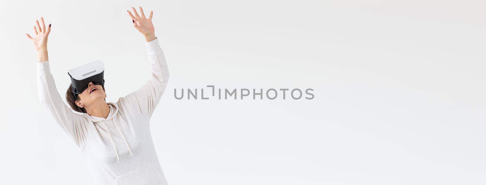 Unidentified middle-aged woman in a light sweater plays a 3D game with glasses of virtual reality on a white background. High technology concept for older people. Advertising space