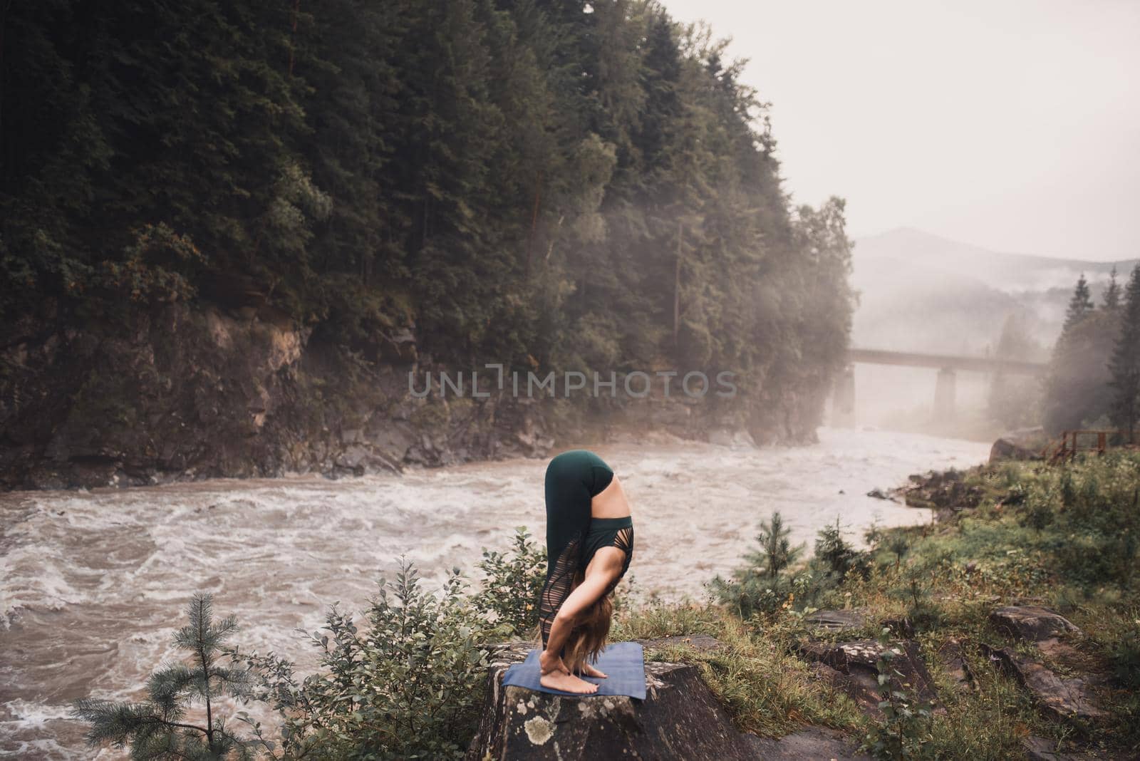 A young blonde woman practices yoga on nature on a stone near a raging river. Green tracksuit.