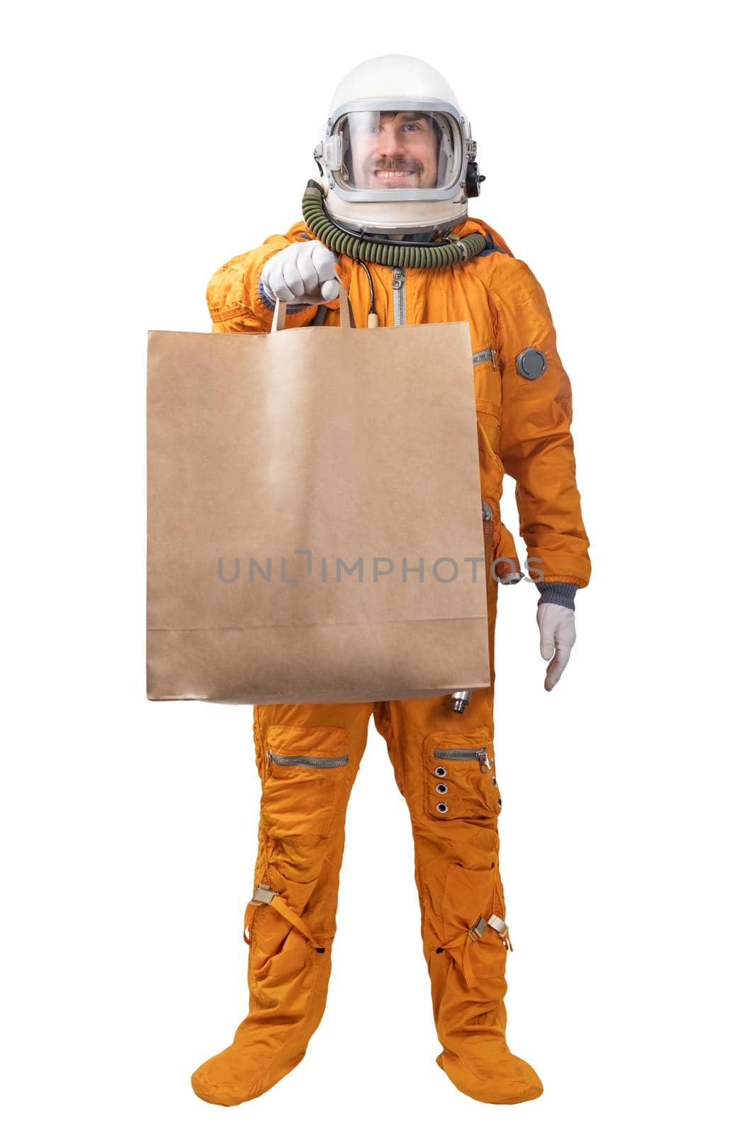 Happy astronaut wearing orange space suit and space helmet holding in hand blank kraft paper bag isolated on white background. Delivery concept. Astronaut courier concept