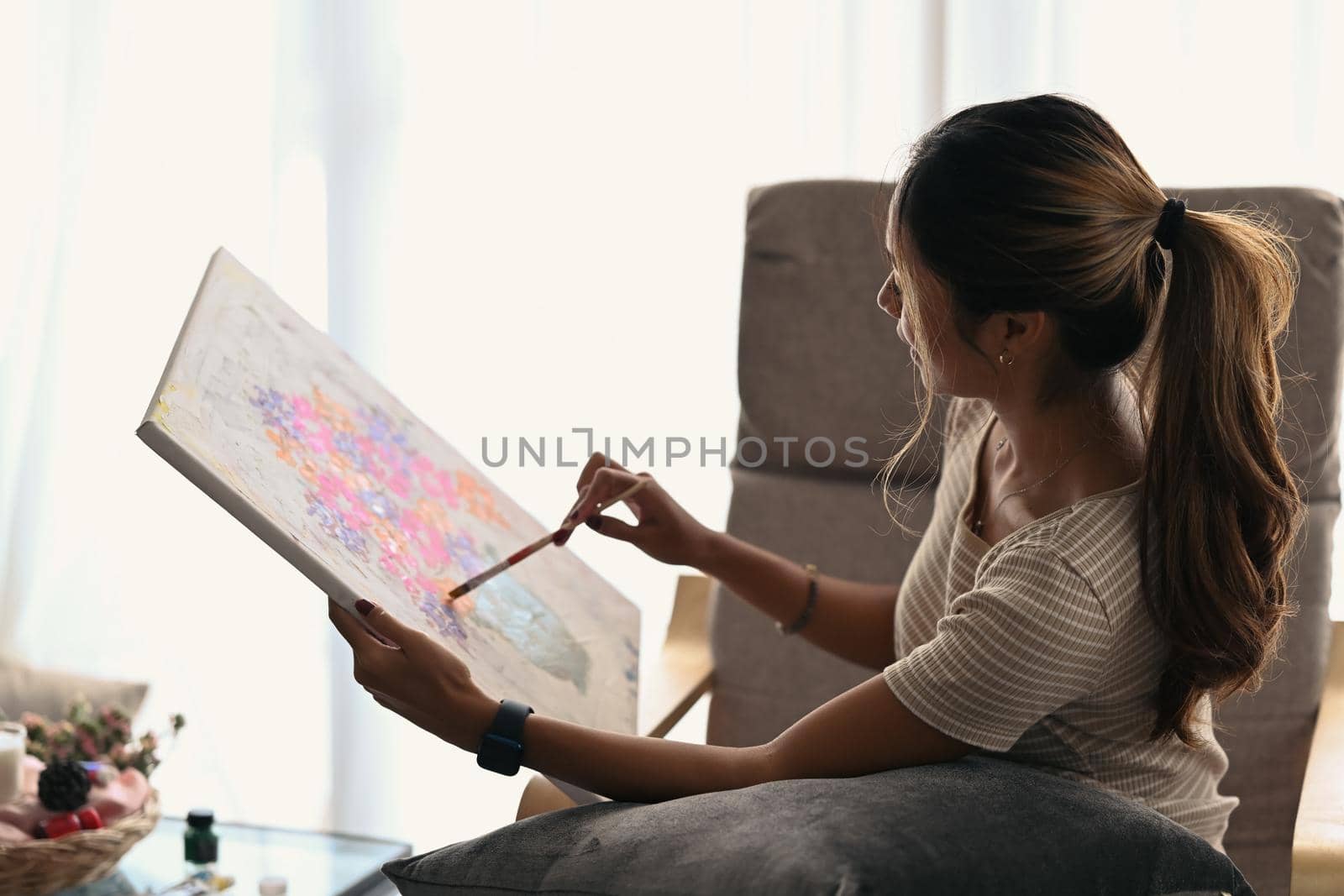 Relaxed young woman sitting on armchair at home and painting on canvas with oil paints.