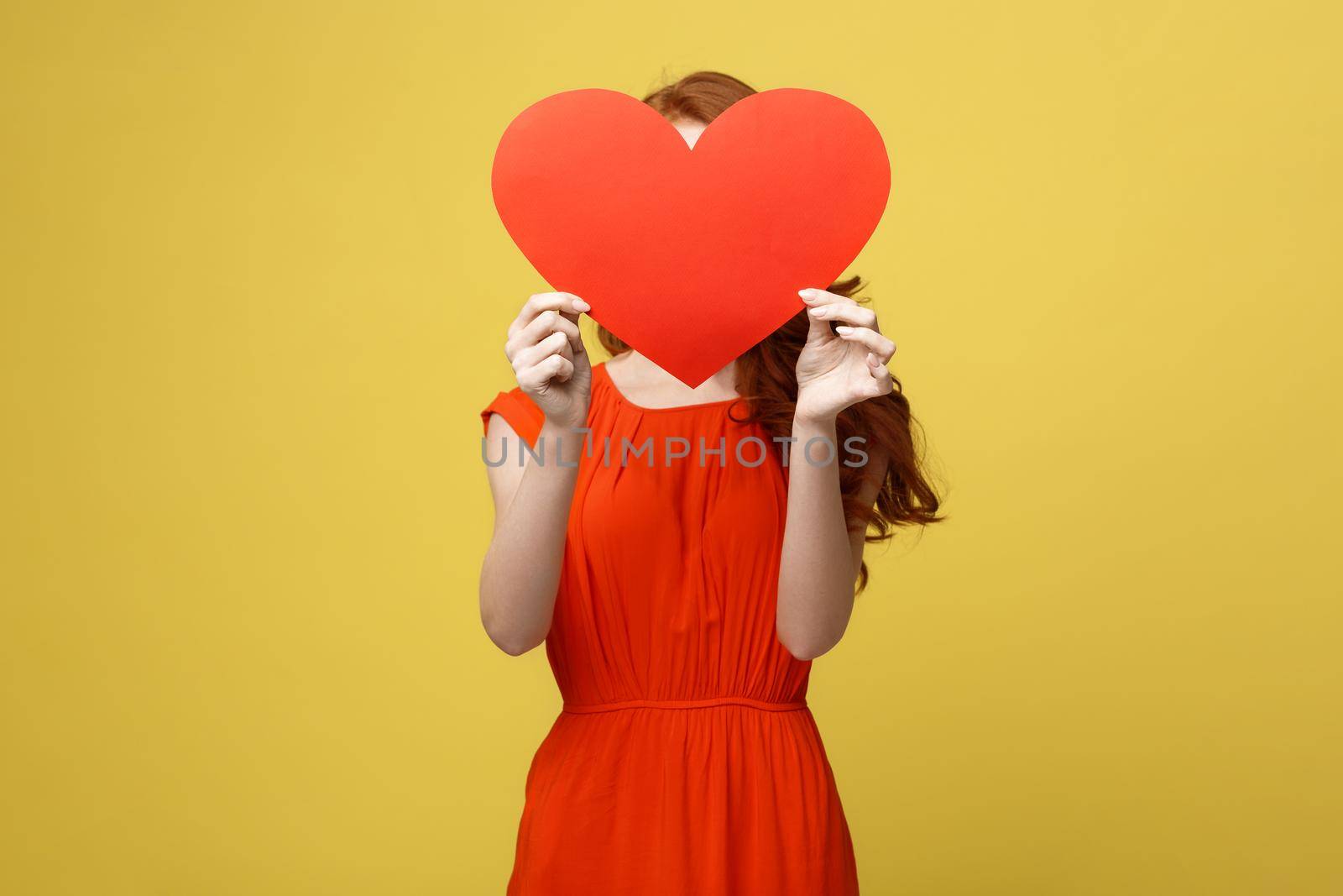 Joyful caucasian girl in orange gorgeous dress holding red heart cover her face. Shallow depth of field. Valentine day concept