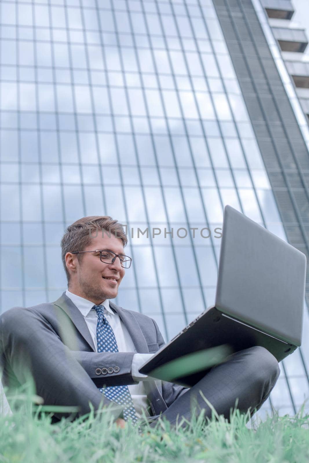 businessman using laptop, sitting on the grass near the office building. photo with copy space