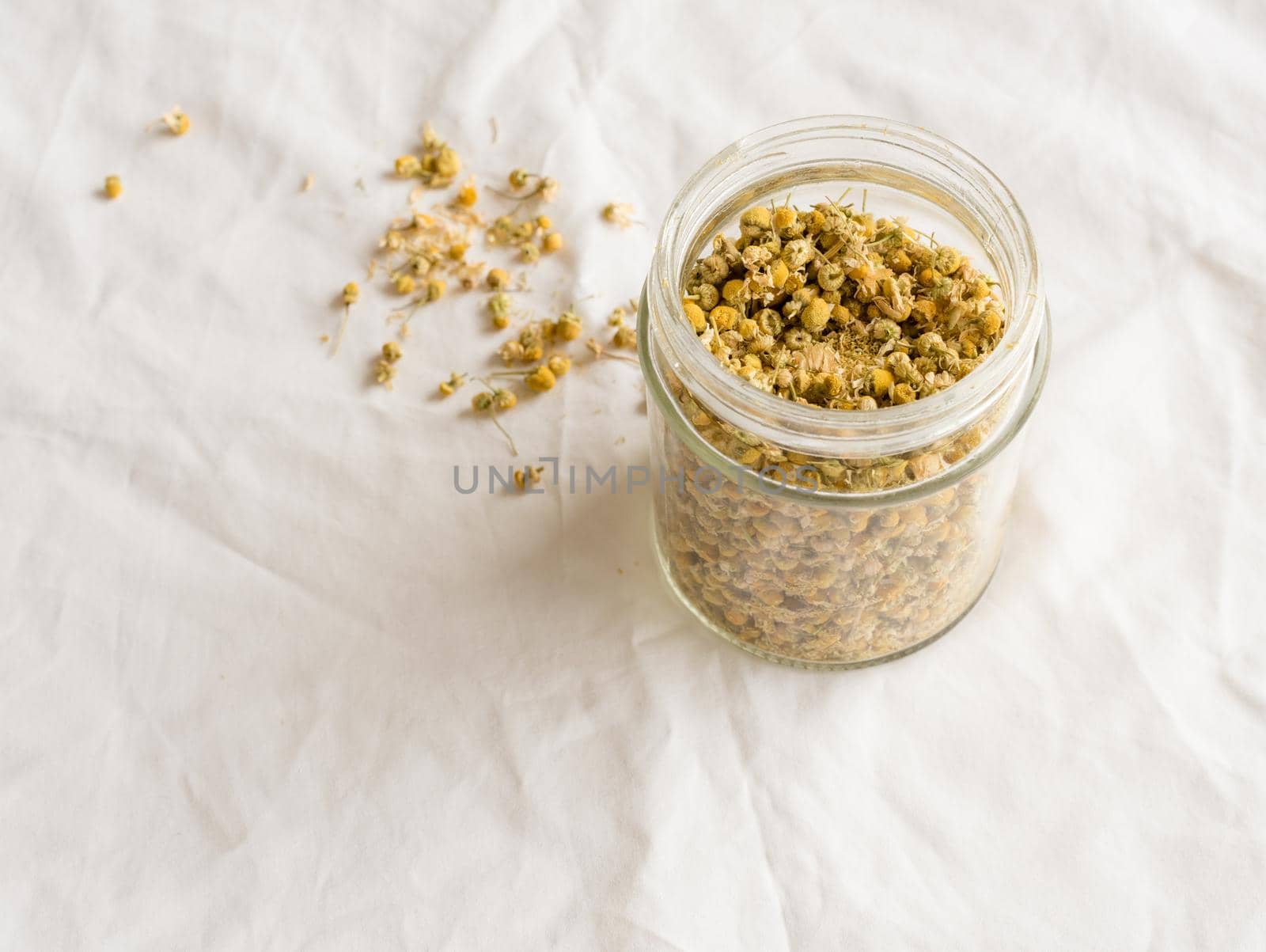 High angle view of dried chamomile flowers for tea in glass jar and scattered on white tablecloth (selective focus)