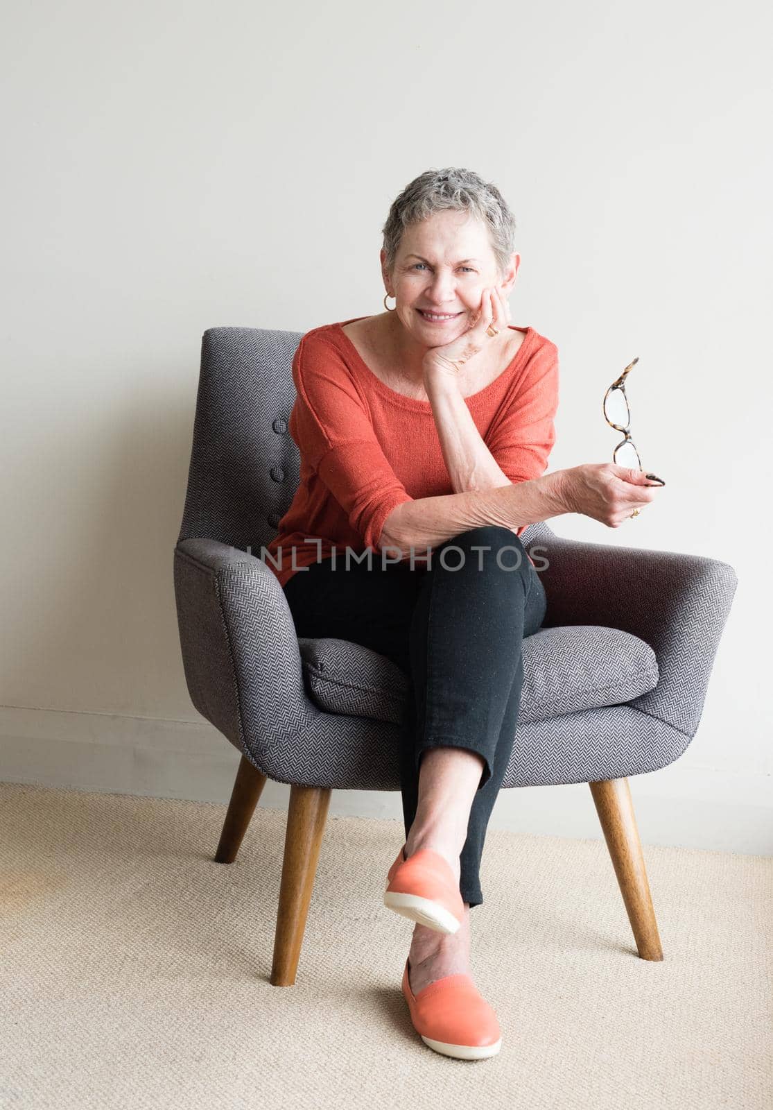 Older woman in orange top seated in armchair holding glasses and smiling