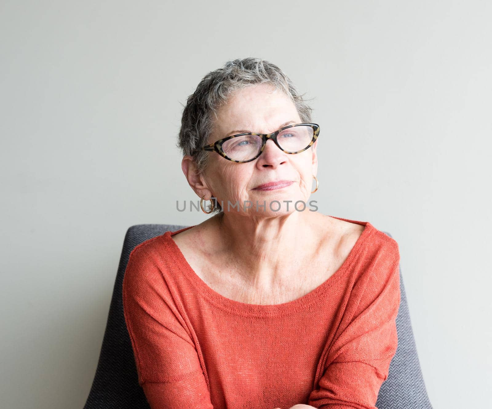 Head and shoulders view of older woman in orange top and glasses seated in armchair against neutral background by natalie_board