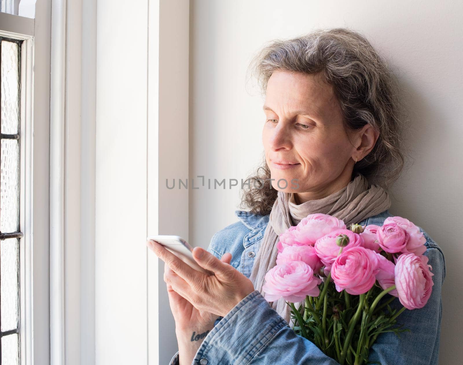 Middle aged woman with grey hair and denim shirt using smart phone, holding pink flowers and smiling by natalie_board