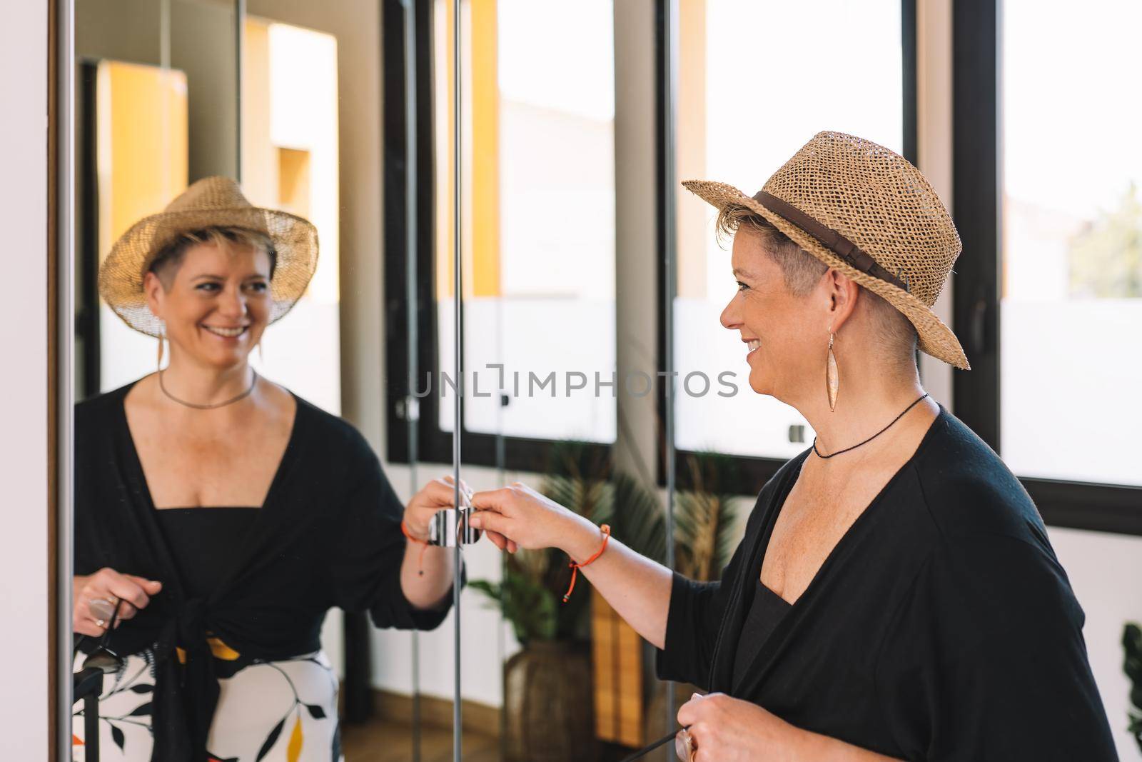 mature woman traveller, looking at herself in the mirror as she dresses to go for a walk along the coast on her summer holiday. bedroom, natural light, dressing room with mirrors.