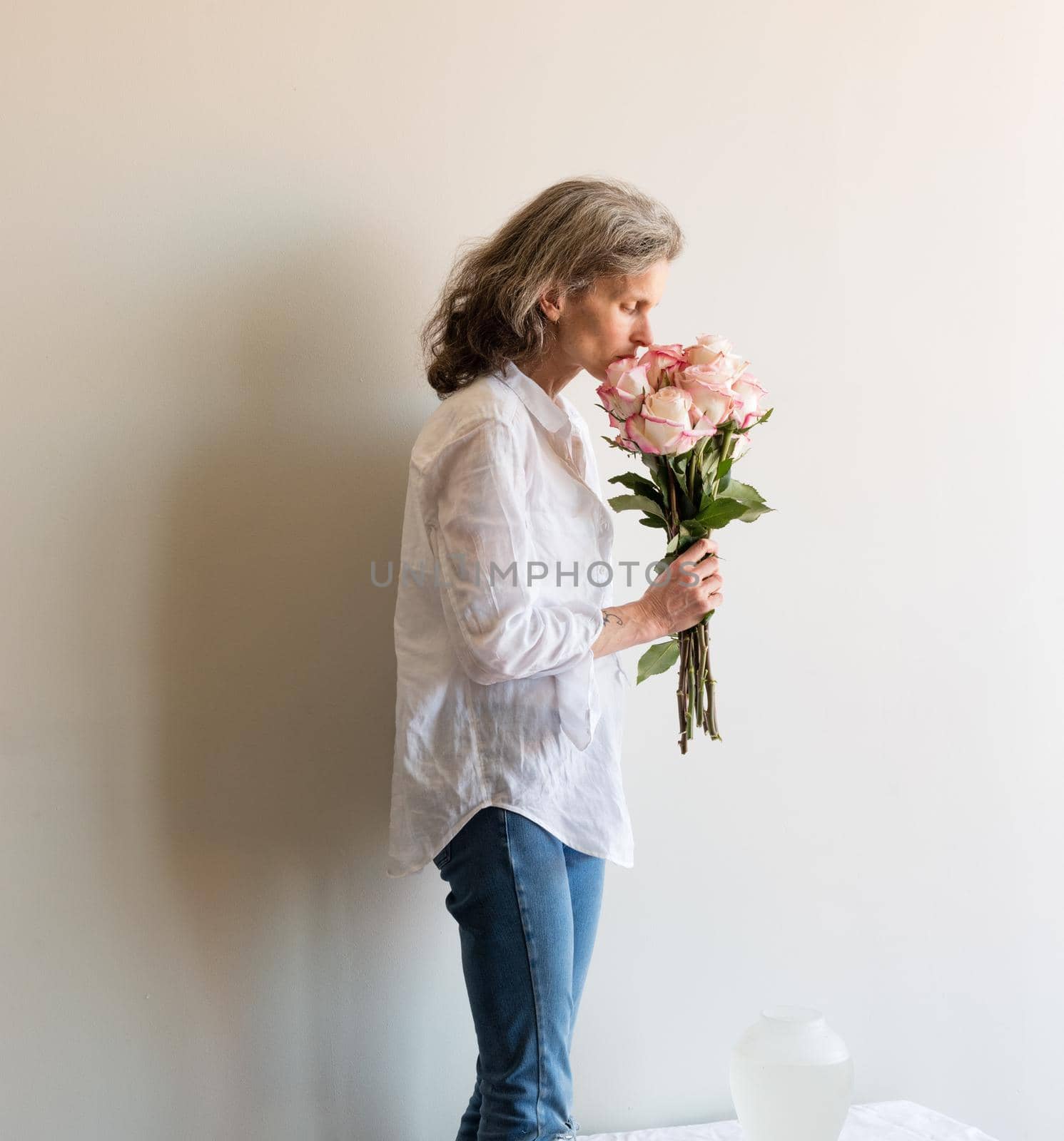 Middle aged woman with grey hair and white shirt smelling pink and cream roses near vase on table