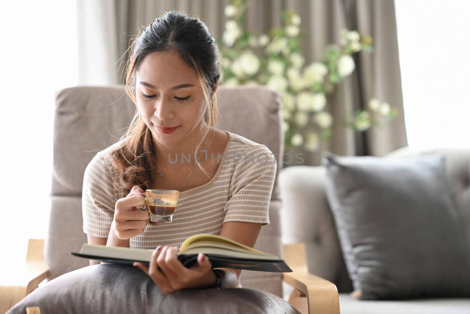 Relaxed calm woman drinking coffee and reading book on comfortable armchair.