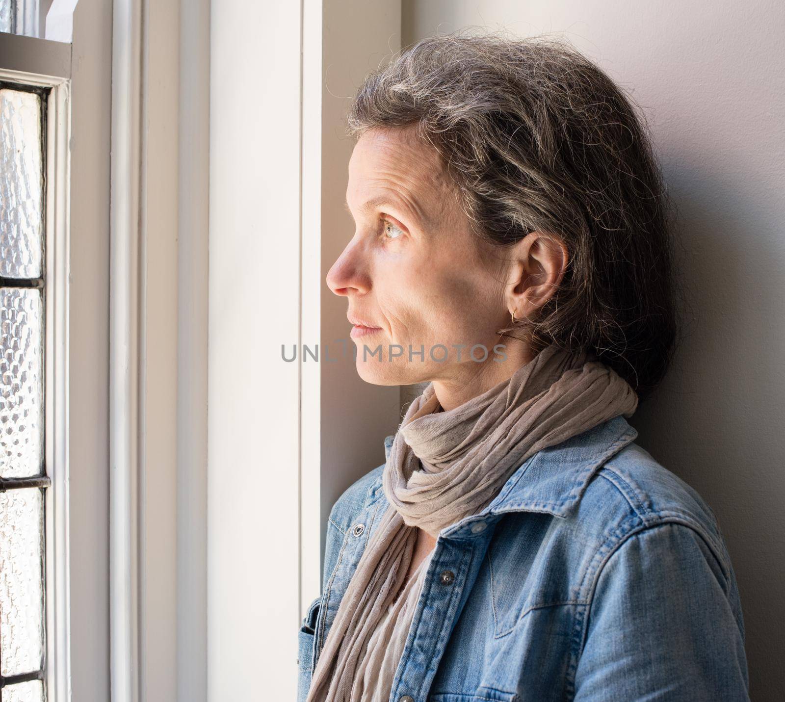 Profile view of middle aged woman with grey hair and scarf and denim shirt next to window looking pensive