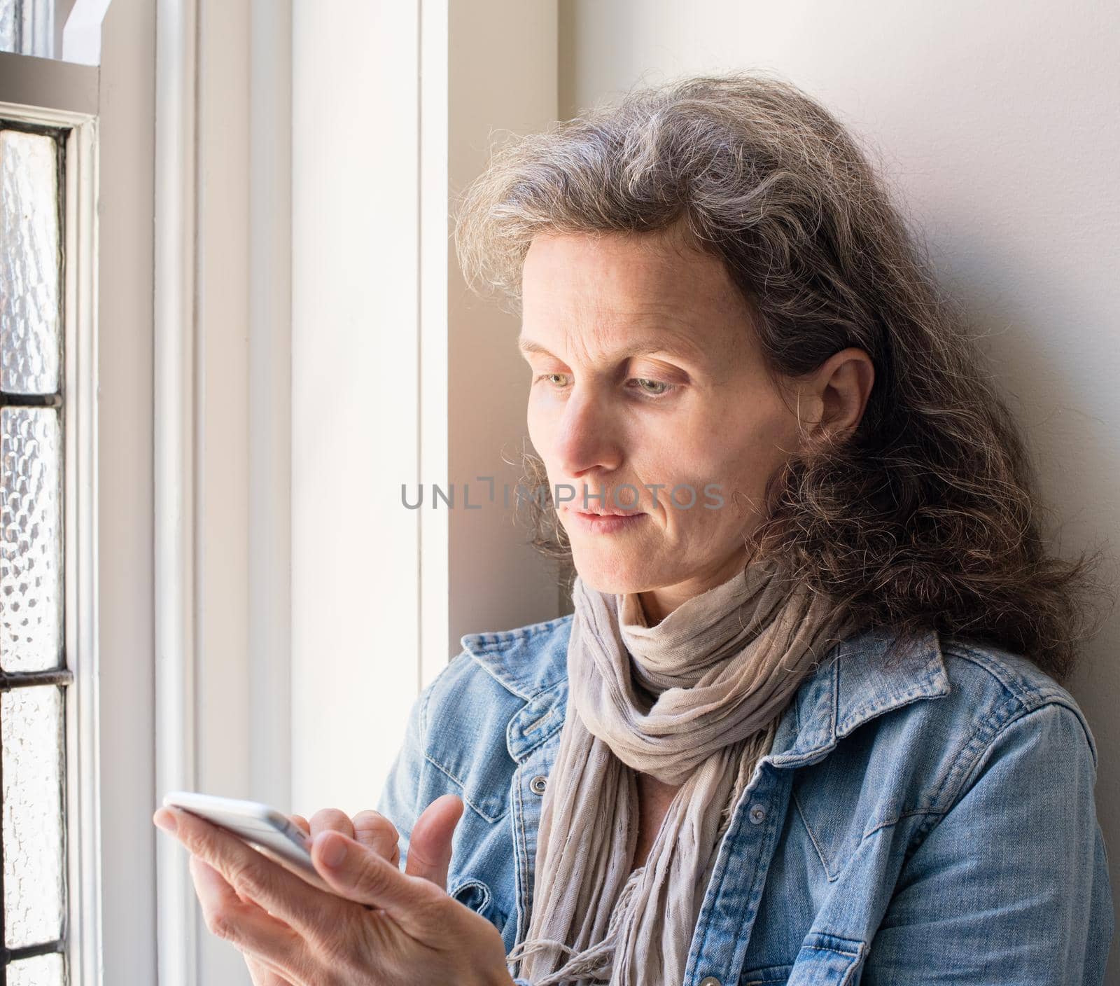 Middle aged woman with grey hair and denim shirt using smart phone next to window (selective focus)