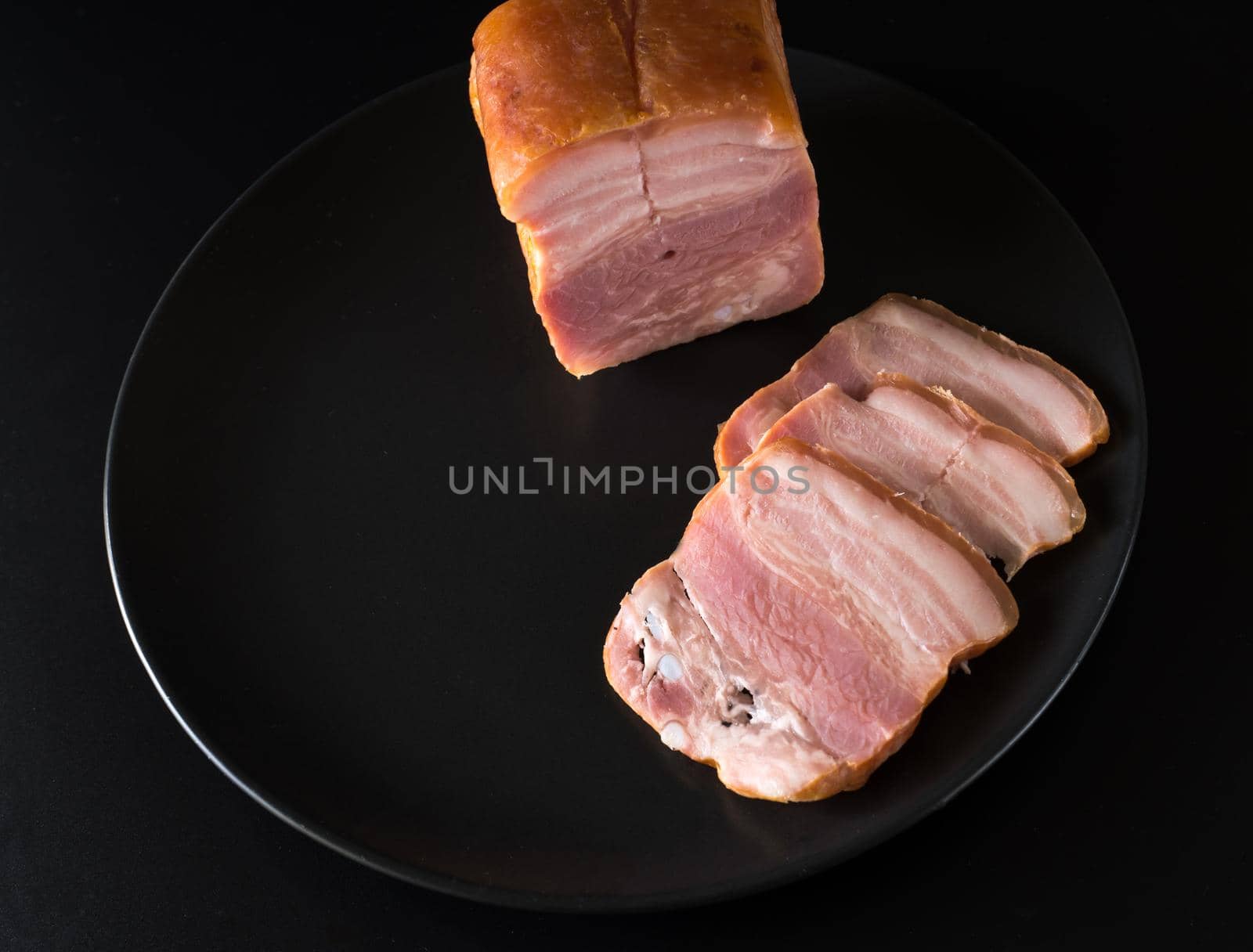 Food, a piece of bacon and cut into slices on a black plate on a black background