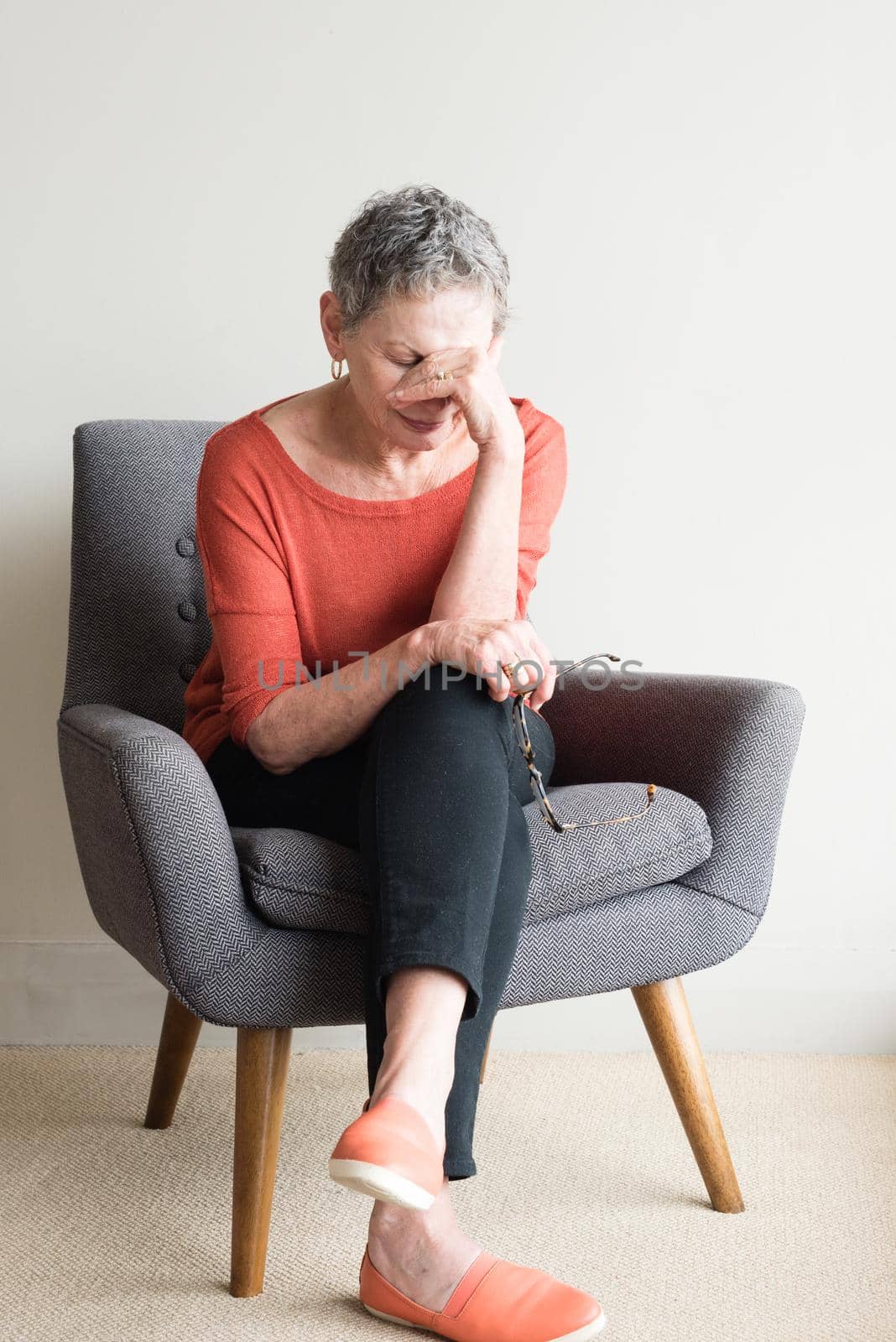 Older woman in orange top seated in armchair with hand over eyes by natalie_board