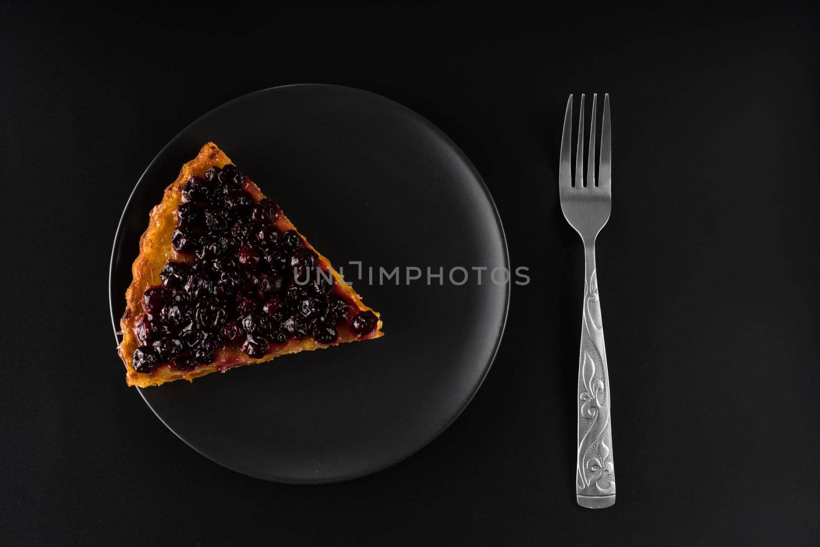 Piece of pie with carrots and currants sprinkled on top on a black plate and a cutlery fork, on a black background