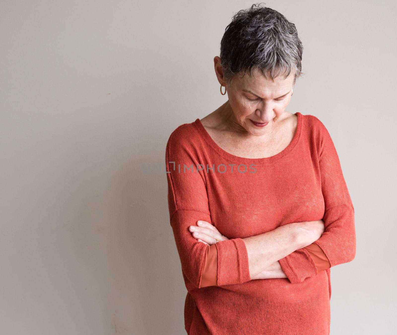 Older woman with short grey hair and orange top looking down pensively with arms crossed