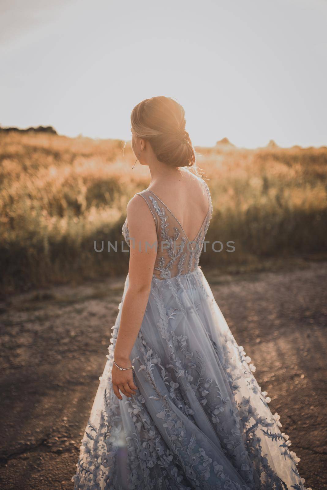 a young girl walks along an asphalt road in a gray blue long dress at sunset. back and arms open. Near the road tall yellow grass grows. The background is blurred.