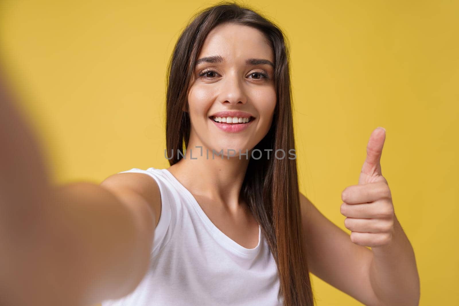 Pleasant attractive girl making selfie in studio and laughing. Good-looking young woman with brown hair taking picture of herself on bright yellow background. by Benzoix