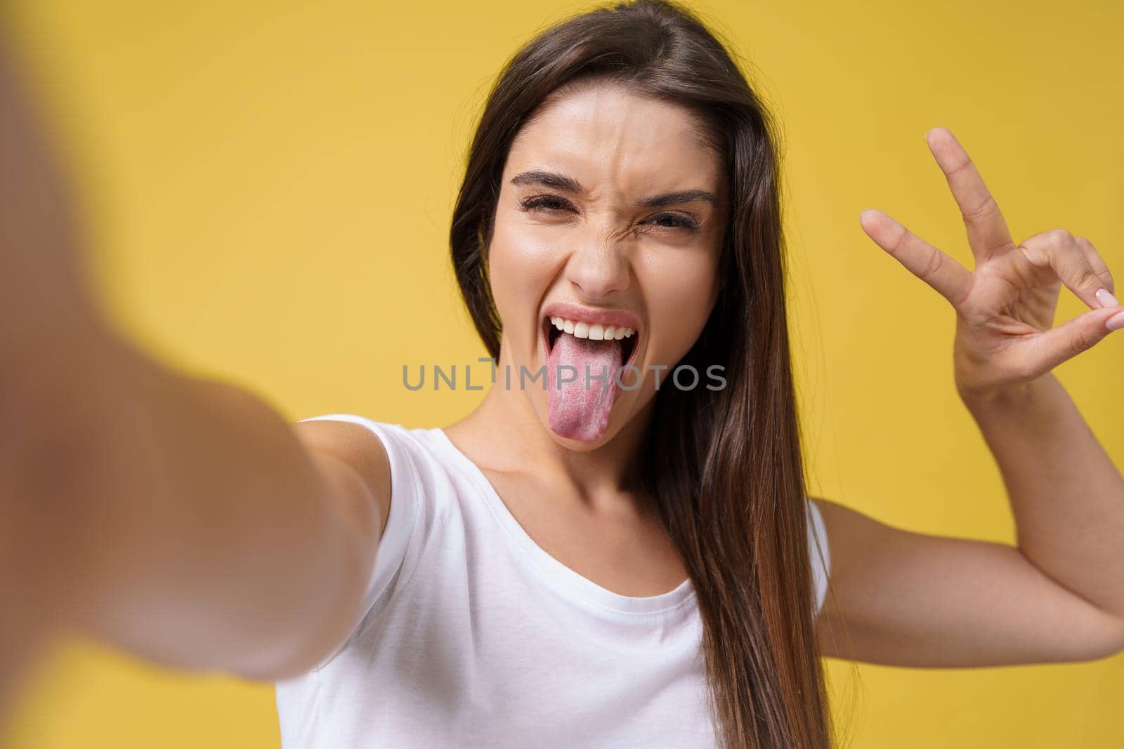 Pleasant attractive girl making selfie in studio and laughing. Good-looking young woman with brown hair taking picture of herself on bright yellow background